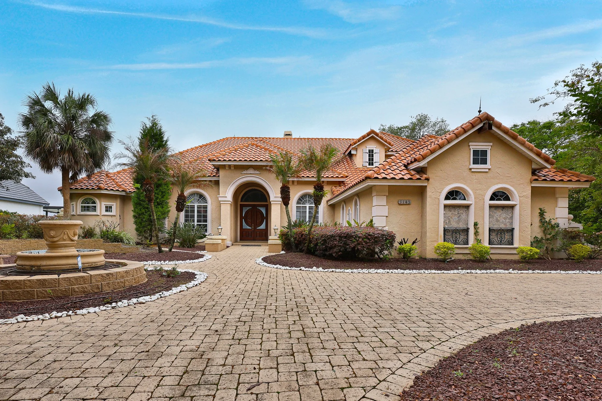 A house with a Spanish-style terracotta-tile roof, arched and rectangular windows, a front door, and a front yard with a curved brick driveway, landscaped with small bushes, palm trees, and a circular fountain with stones surrounding it.