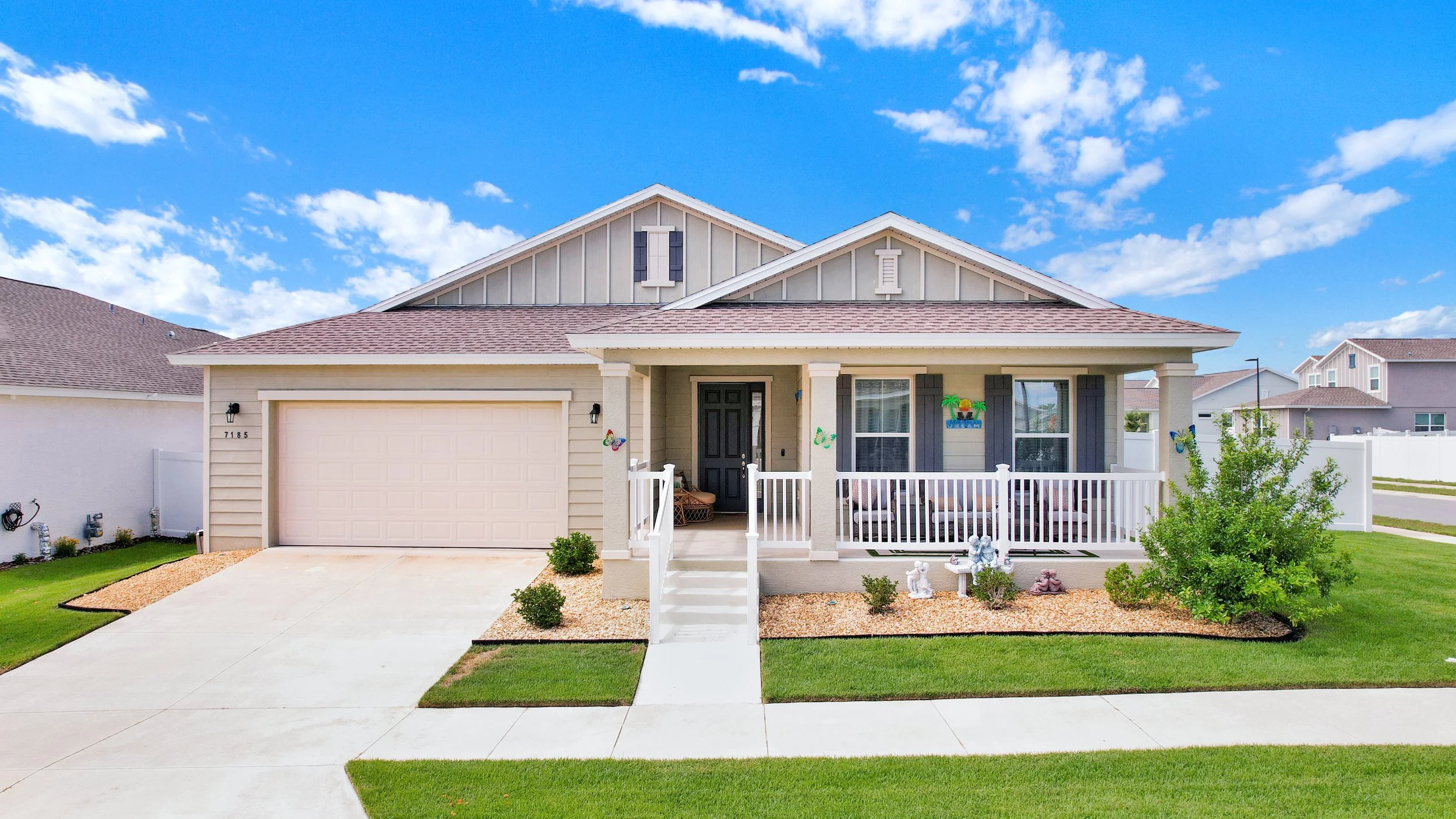 Front view of a modern suburban house with a two-car garage, white porch railing, and decorated yard, under a bright blue sky with scattered clouds.