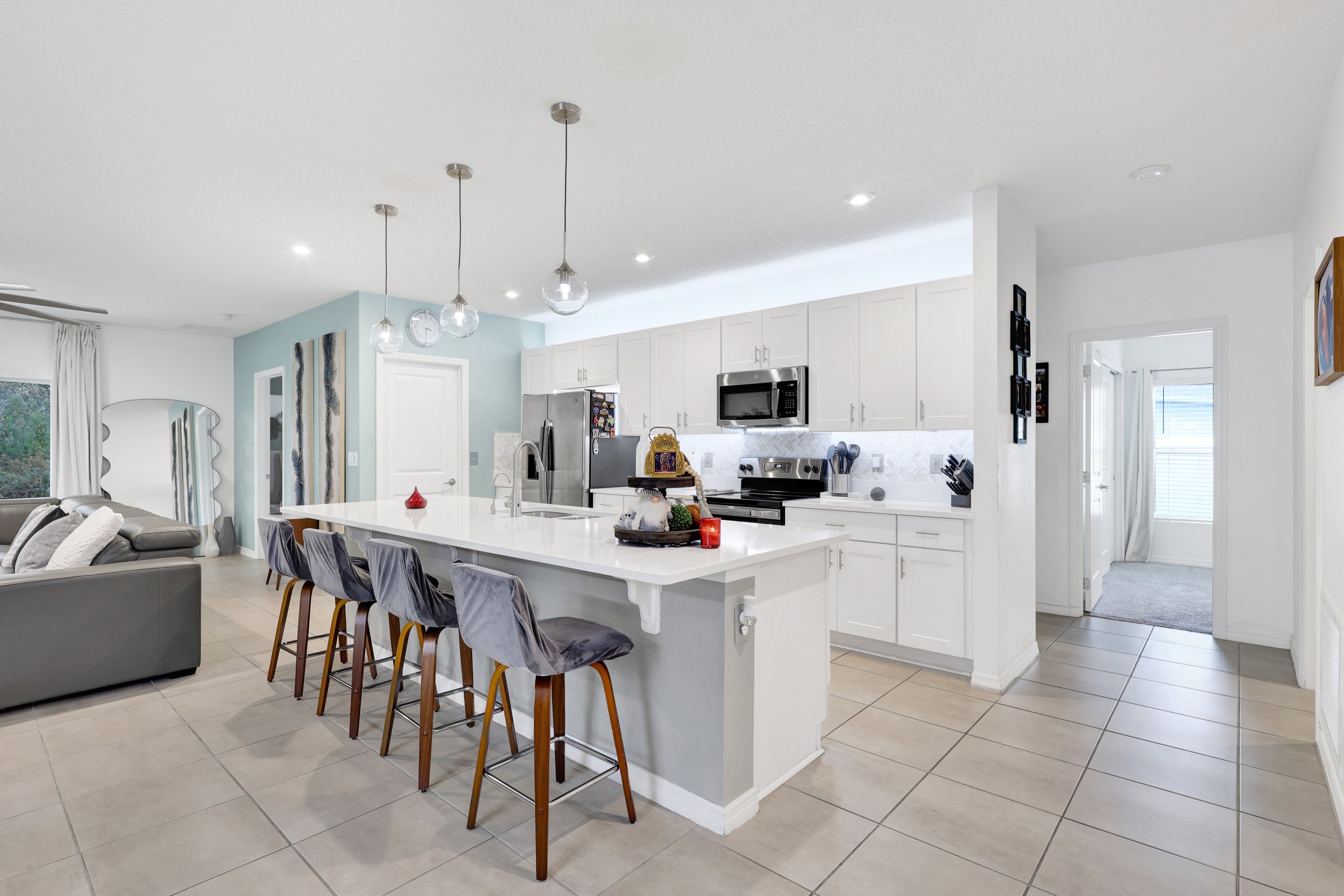 Open-concept kitchen and living area with white cabinets, stainless steel appliances, a center island, and modern pendant lighting. A dining table with upholstered chairs is also visible.