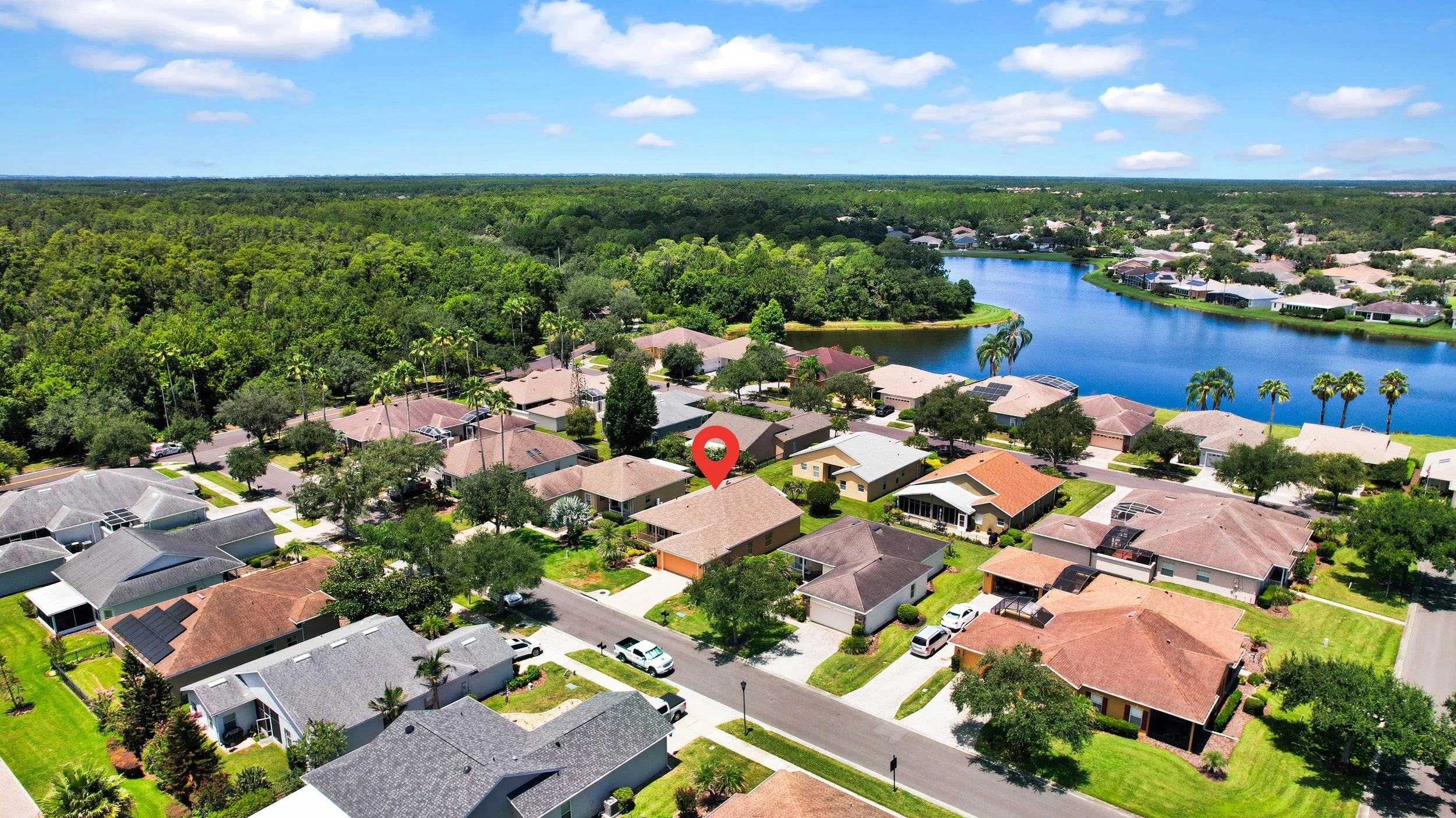 Aerial view of a suburban neighborhood with houses, a pond, and green trees under a partly cloudy sky.