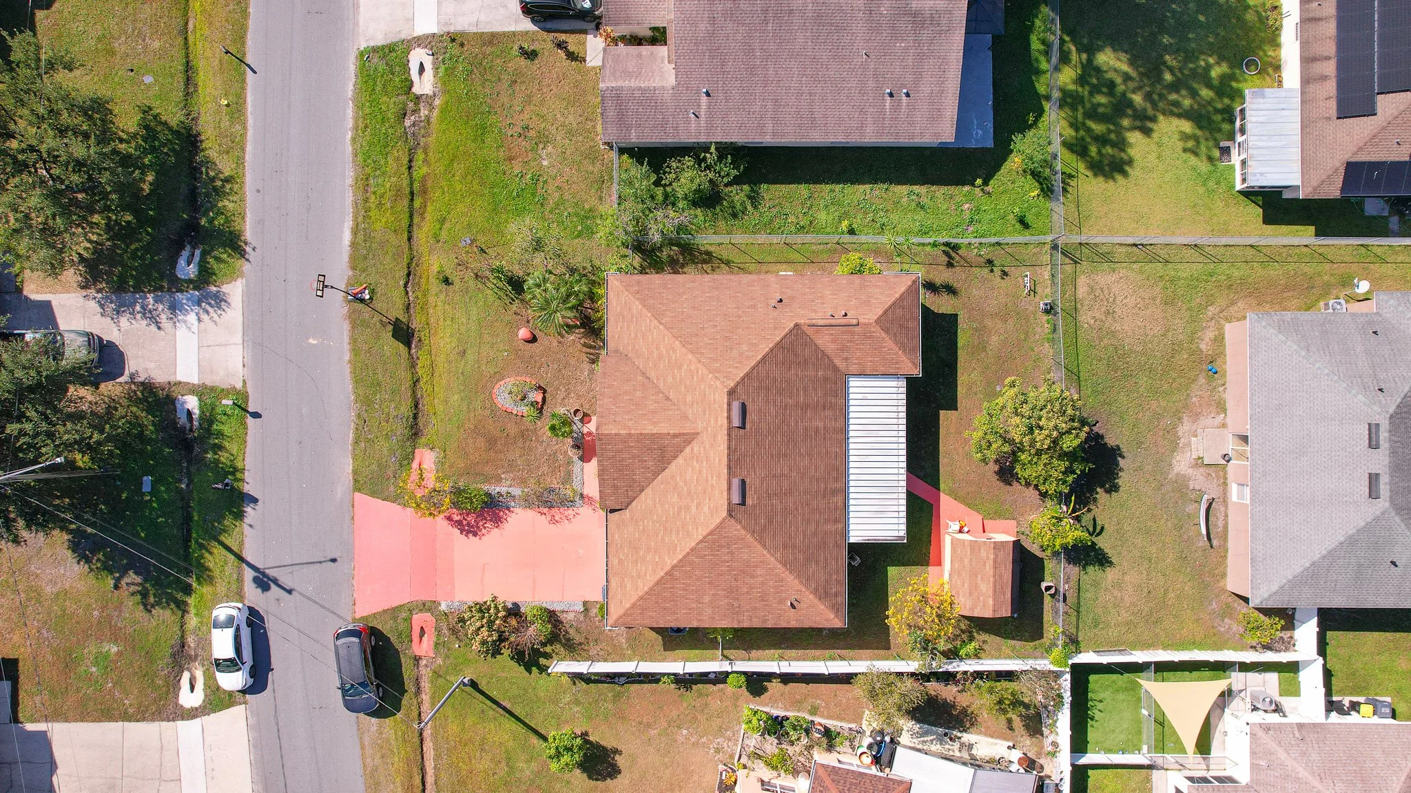 An aerial view of residential houses, streets, and yards in a neighborhood with green lawns, trees, parked cars, and basketball hoops.