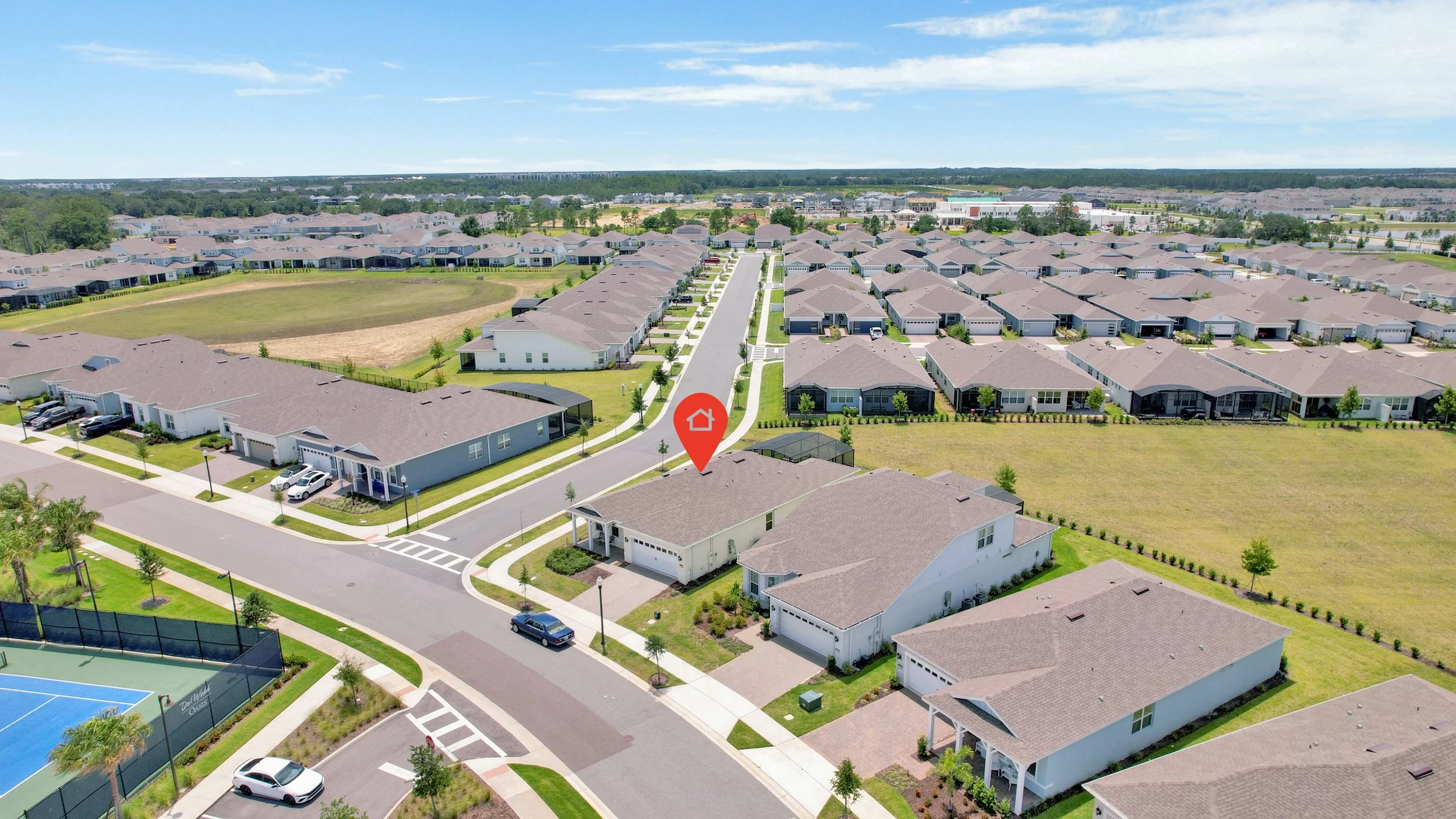 Aerial view of a suburban neighborhood with rows of single-family homes, green lawns, and a park area, under a blue sky with scattered clouds.