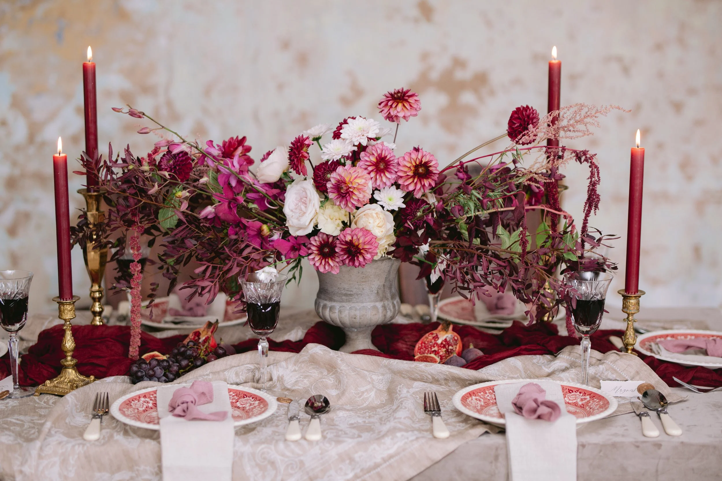 Ein festlich gedeckter Tisch mit einer großen Blumenarrangement in der Mitte, vier rote Kerzen, Wein- und Wassergläser, Teller mit rotem Muster, Besteck, Lichtblumen, Trauben und Feigen, sowie Platzkarten.