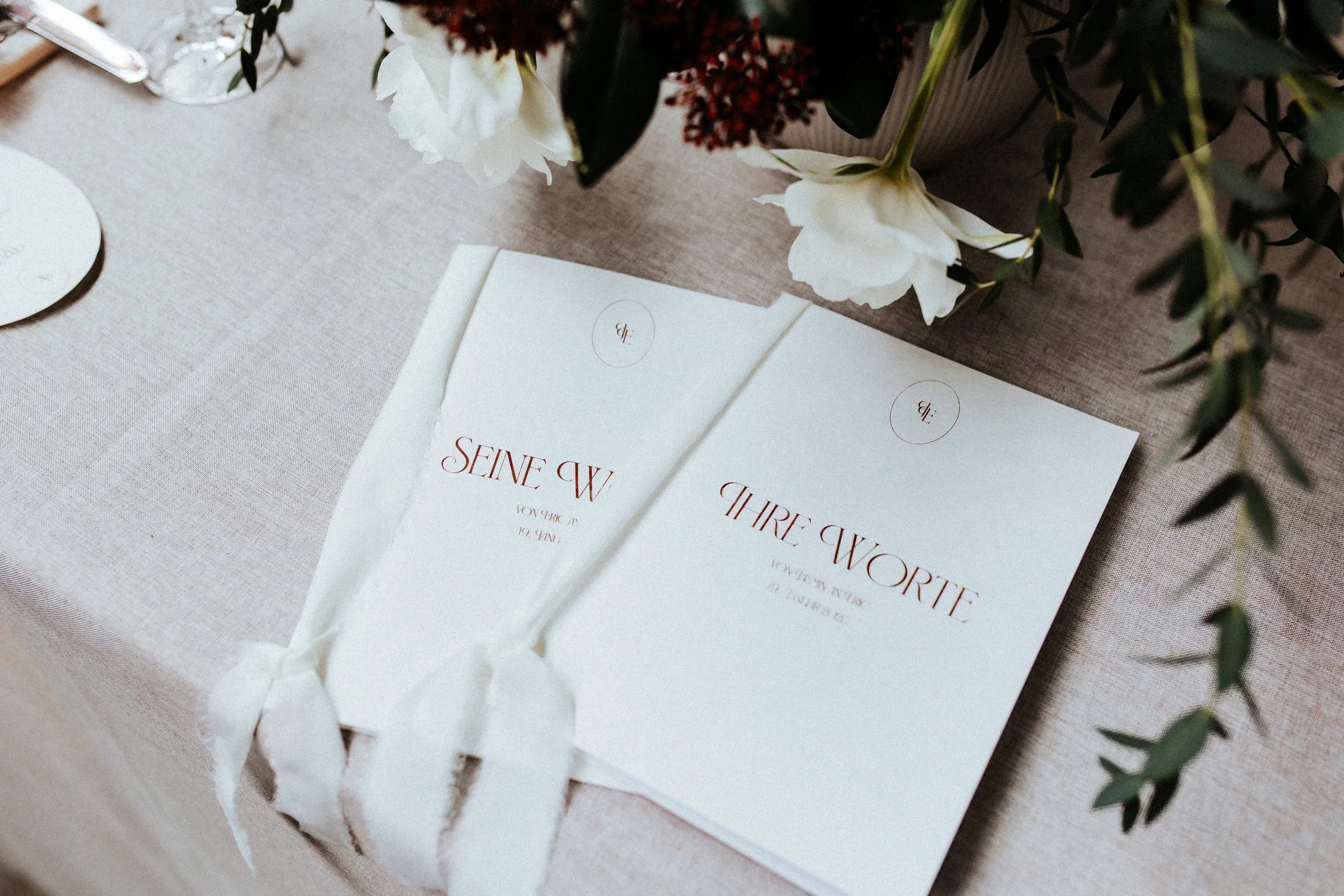 Zwei weiße Karten mit roten und grauen Texten, die 'Seine Worte' und 'Ihre Worte' heißen, liegen auf einem beige Stofftisch. Eine Blumenarrangement mit weißen und roten Blumen befindet sich darüber. Das zeigt die Papeterie zur Trauung.