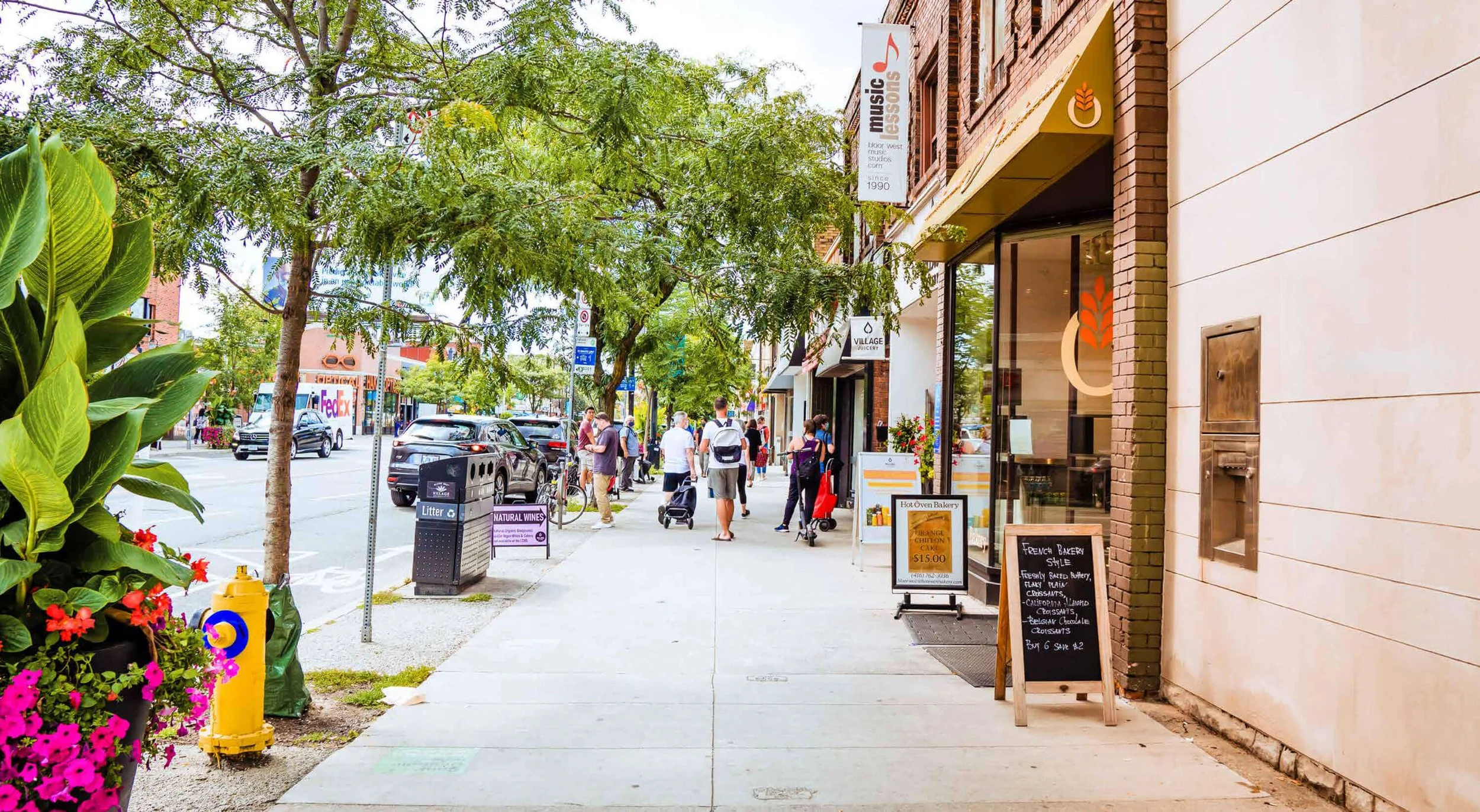 City sidewalk with businesses, pedestrians, and parked cars, including flower planters, trees, street signs, and a menu board outside a bakery.
