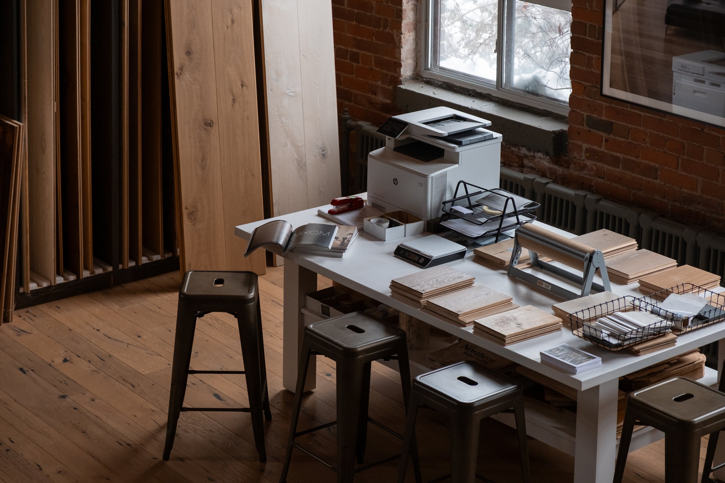 Interior of an office or studio with a white table holding a printer, stacks of paper, and office supplies. There are three black stools around the table, wooden floor, and a large window with a brick wall outside.