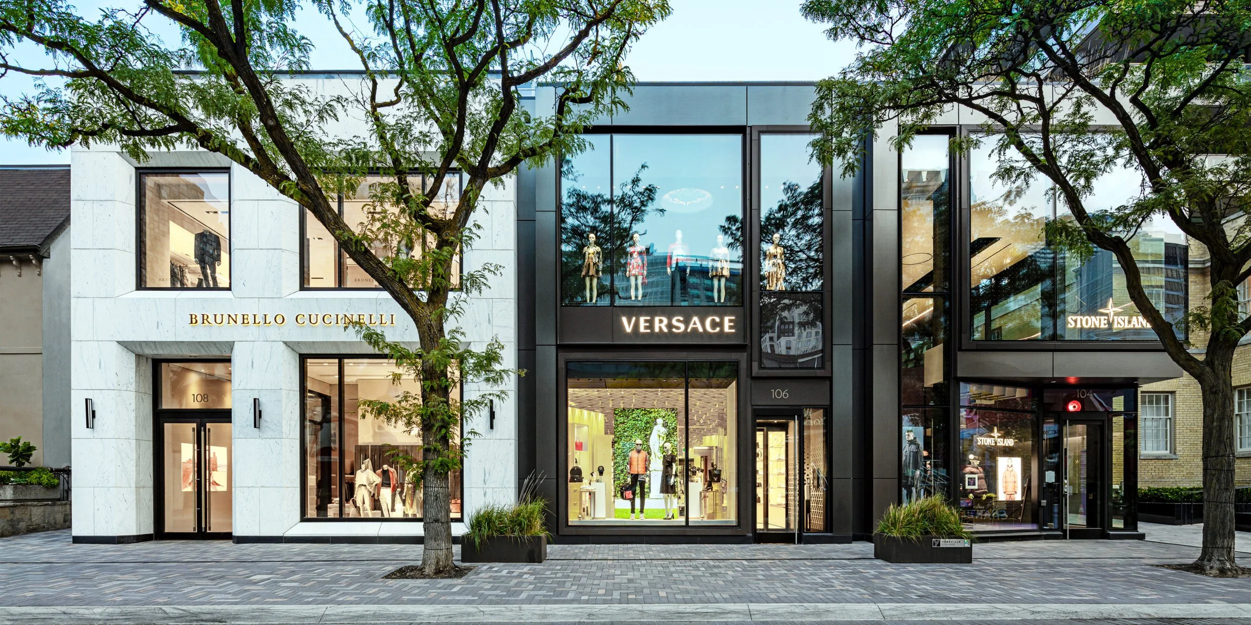 A modern retail storefront with three luxury brands: Brunello Cucinelli, Versace, and Stone Island, visible signage, mannequins in the window, and trees in the foreground.