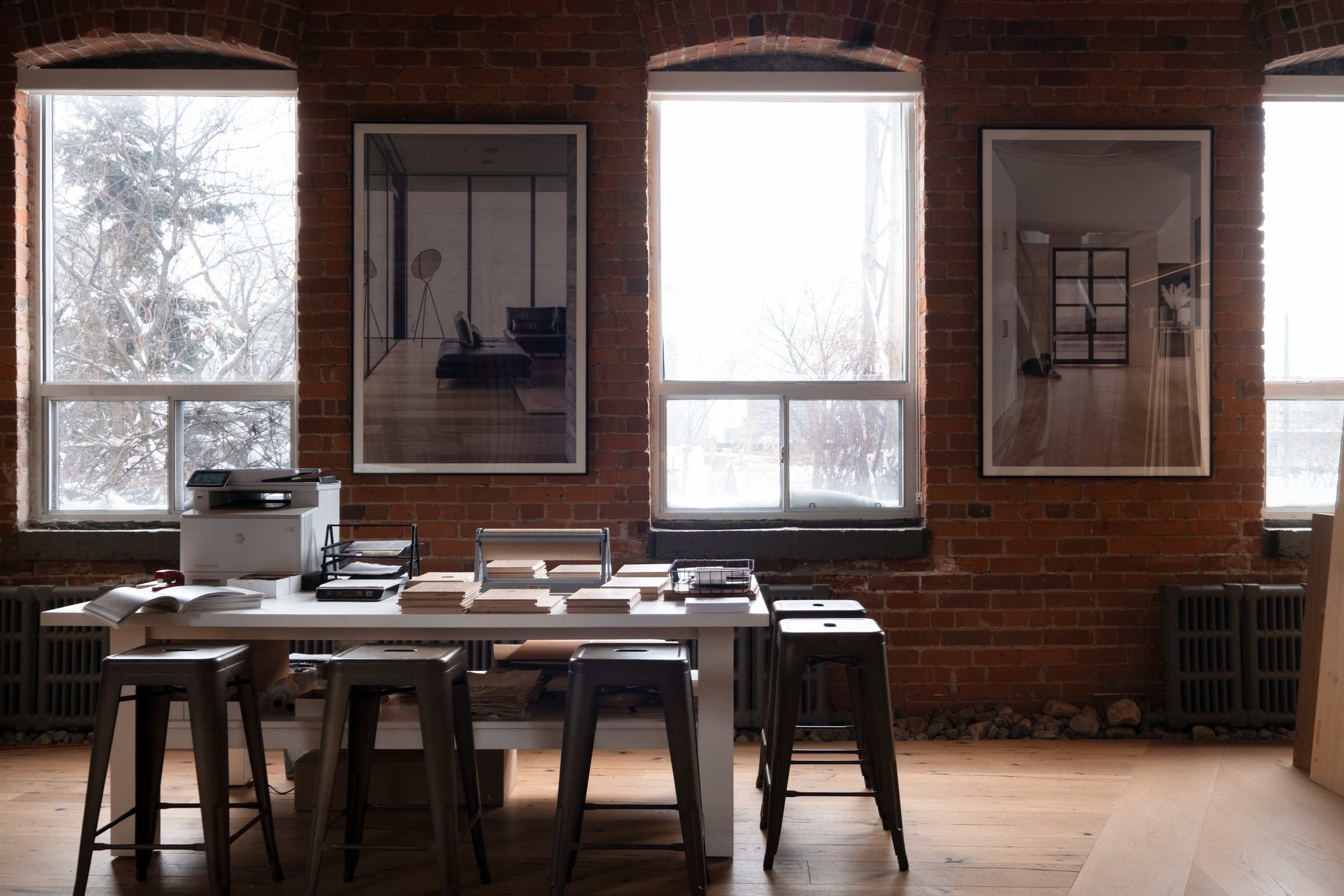 Interior of an office space with a brick wall, three large windows, a white table with stacks of papers, a printer, and five black bar stools.