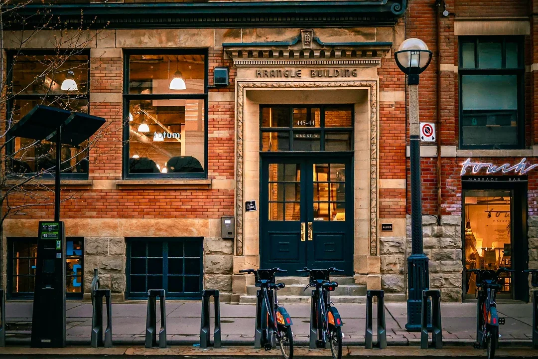 A brick building labeled 'Kangle Building' with large windows and a dark green door, bicycles parked in front, and a streetlamp on the sidewalk.