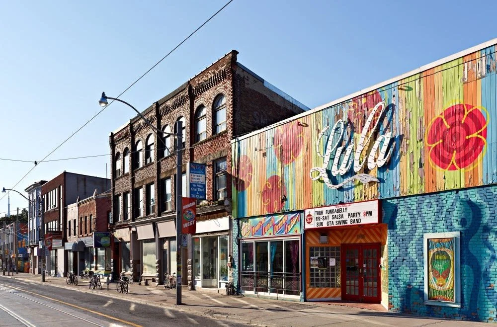 Street view of a colorful building with a large painted sign that says "Lolla." The building has a mural with abstract patterns and flowers, and an electronic marquee advertising events like salsa parties and a swing band. Bicycles are parked along the sidewalk under streetlights, with additional storefronts visible along the street.