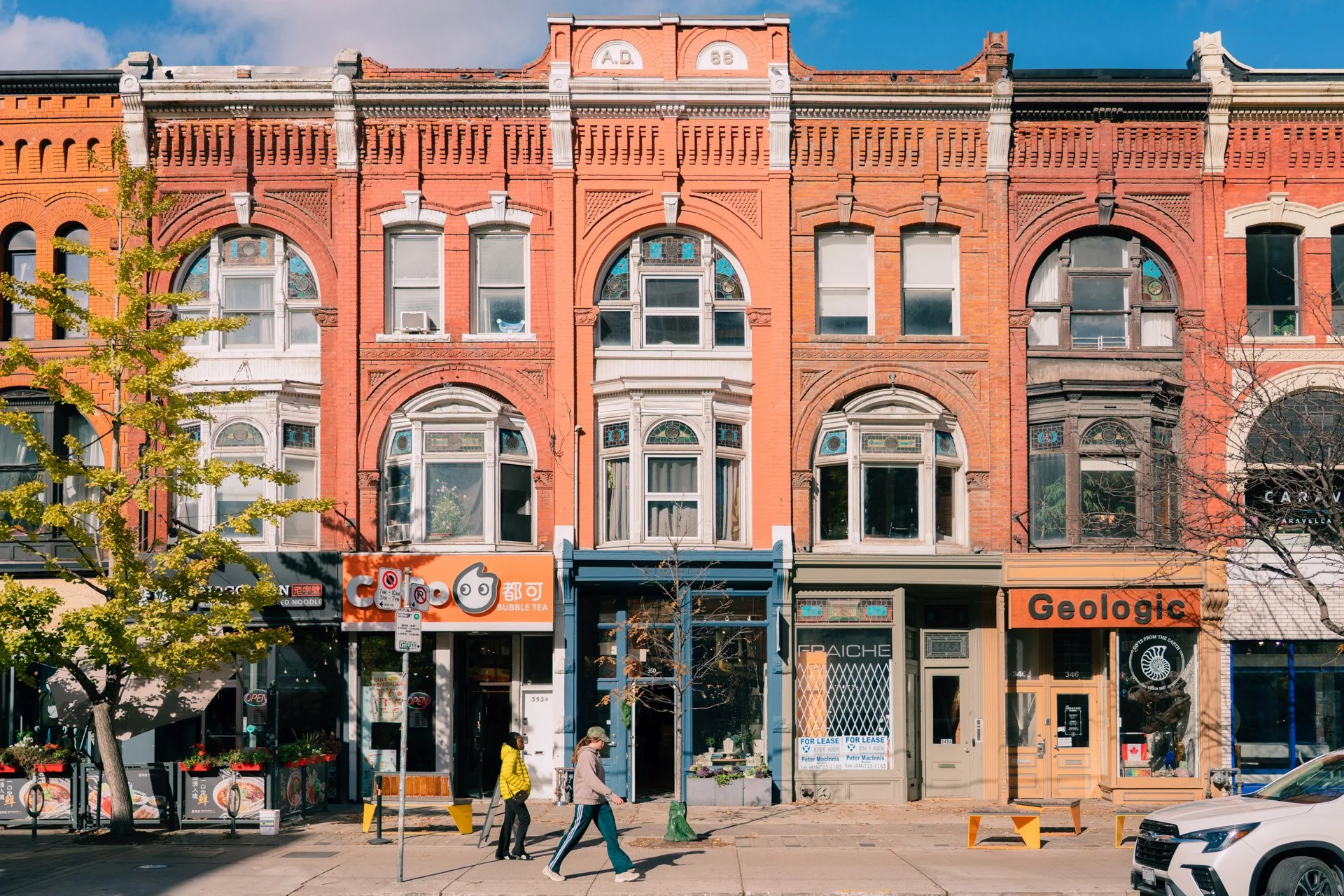 A row of historic red brick buildings with decorative architectural details and storefronts on the ground floor, including a bubble tea shop and other businesses, with pedestrians walking on the sidewalk and a tree in the foreground.