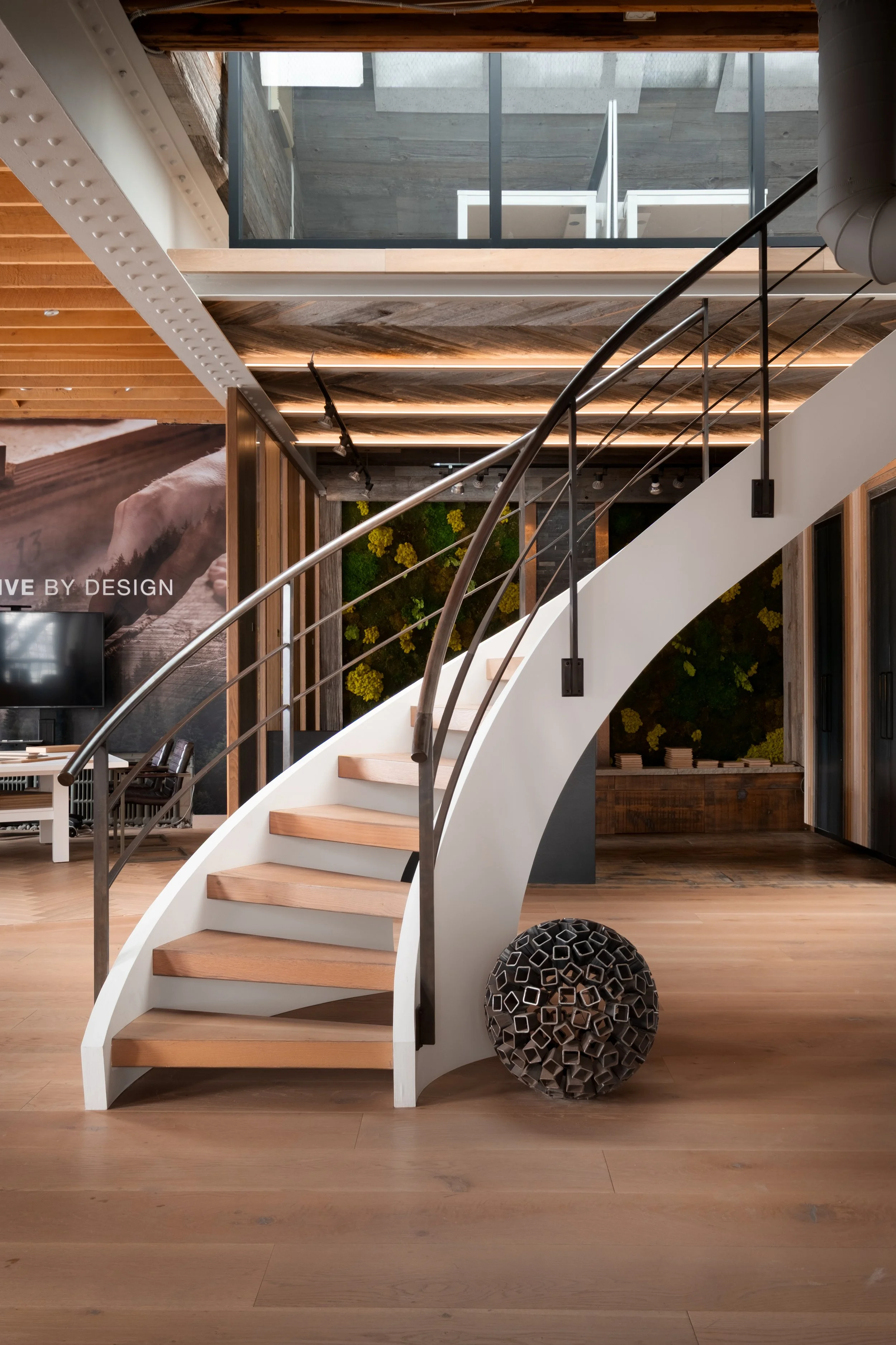 Interior of a modern, stylish building with a curved staircase featuring wooden steps, black metal railing, and a spherical decorative object at the base of the stairs. Visible upper floor and green wall with plants in the background.