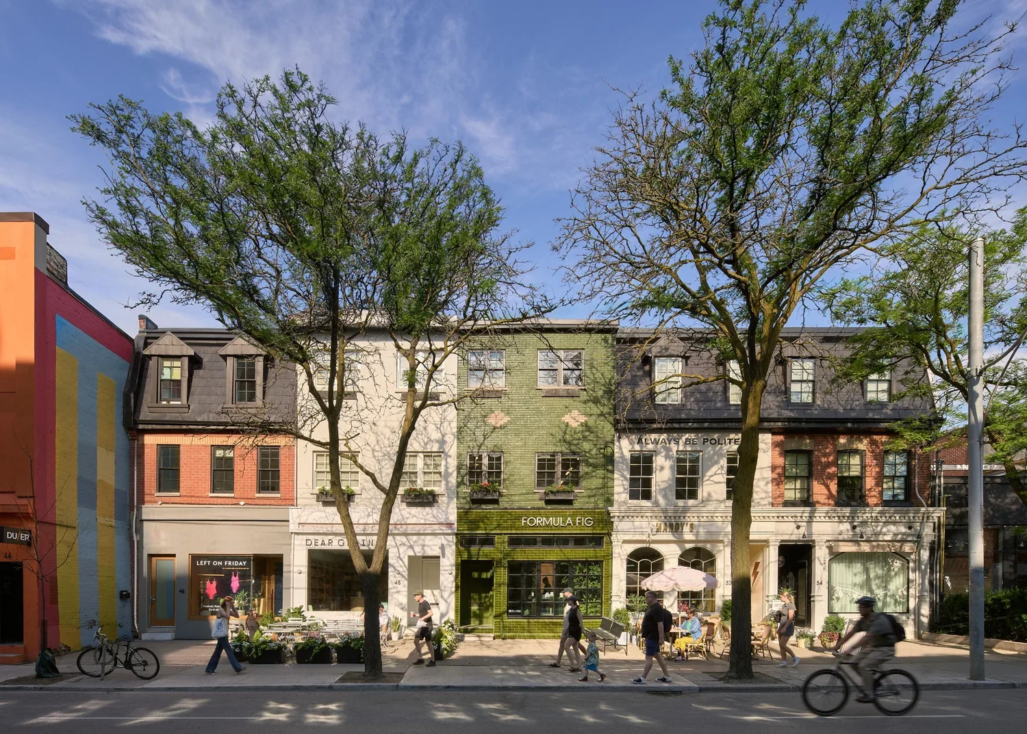 Street view showing a row of small shops with people walking and riding bicycles on a tree-lined sidewalk, under a partly cloudy sky.