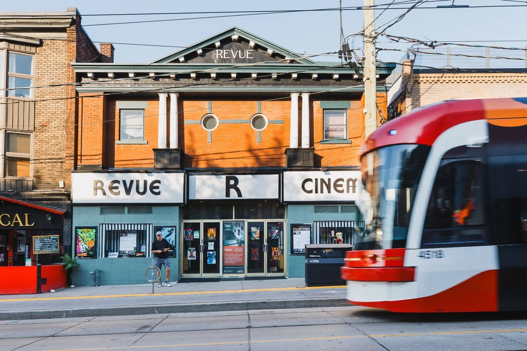 The Revue Cinema building with a marquee sign and a man walking on the sidewalk in front, while a red and white bus passes by.