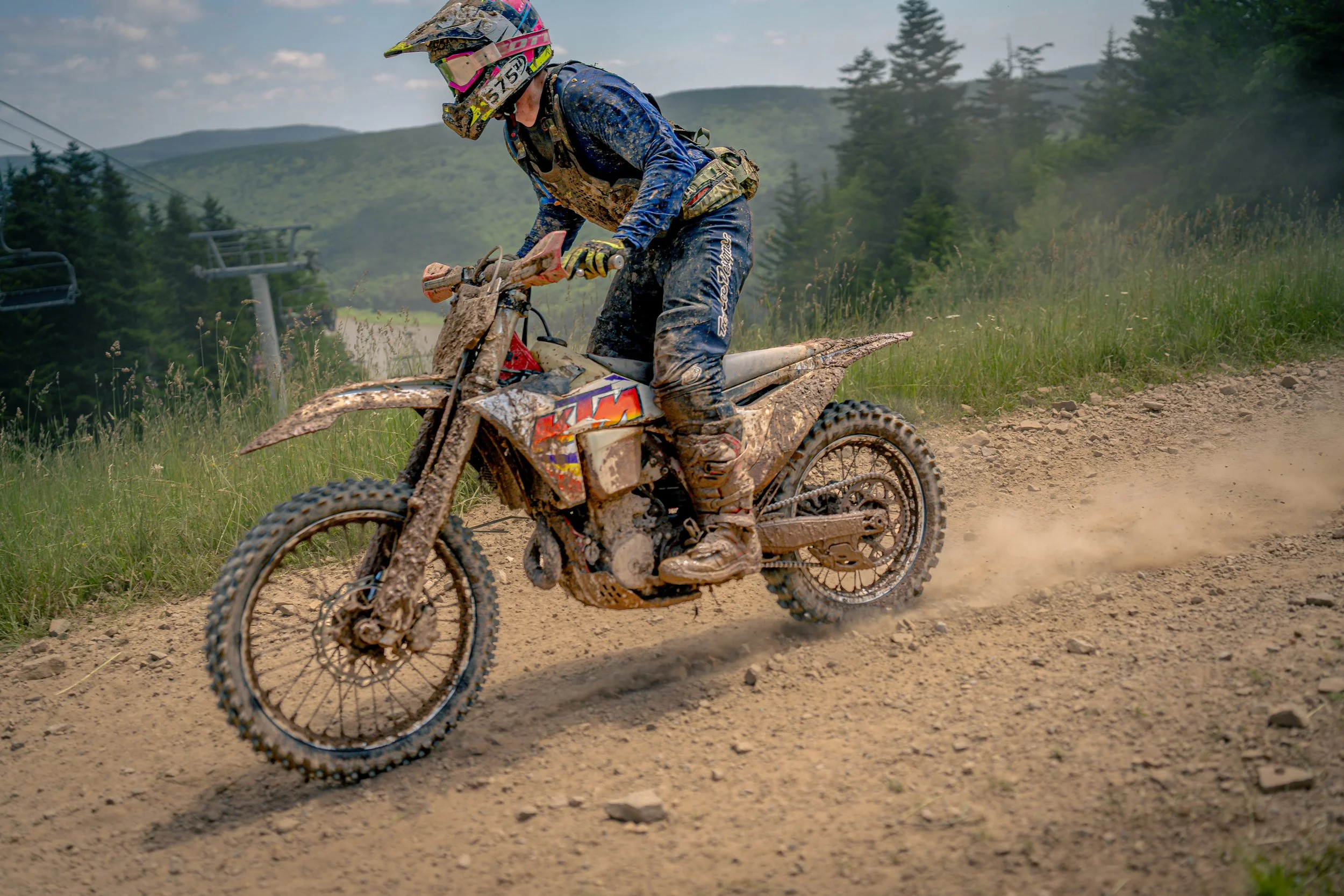 A motocross rider in blue gear and a helmet riding a muddy dirt bike on a trail in a forested mountainous area.