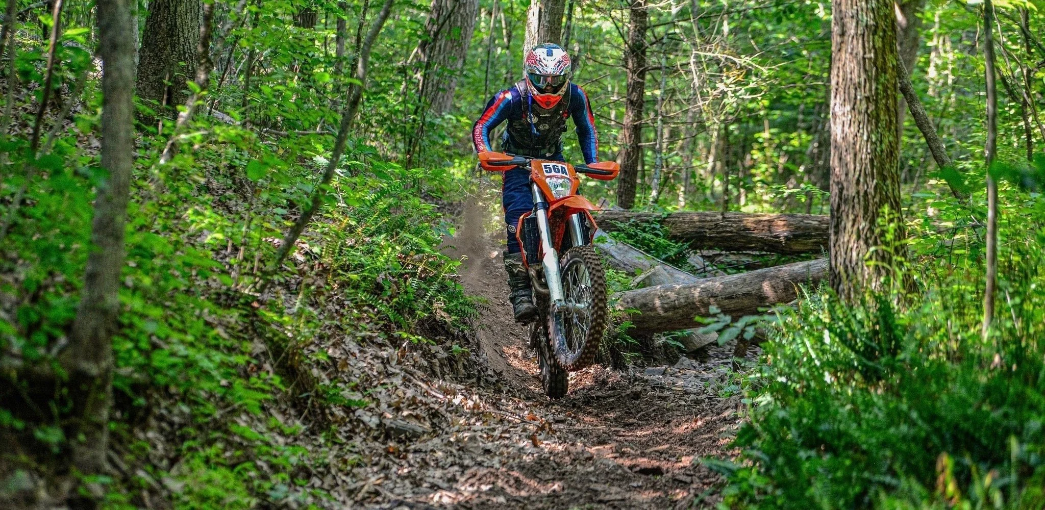 A person riding a dirt bike on a forest trail, wearing a helmet, goggles, gloves, and protective gear, with trees and greenery surrounding the trail.