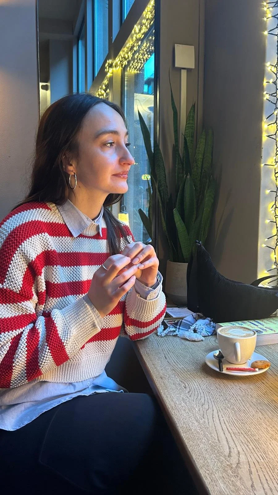 A woman with dark brown hair, wearing a red and white striped sweater, sitting at a wooden table in a cozy cafe by a window with decorative string lights, enjoying a hot beverage and holding a cookie.