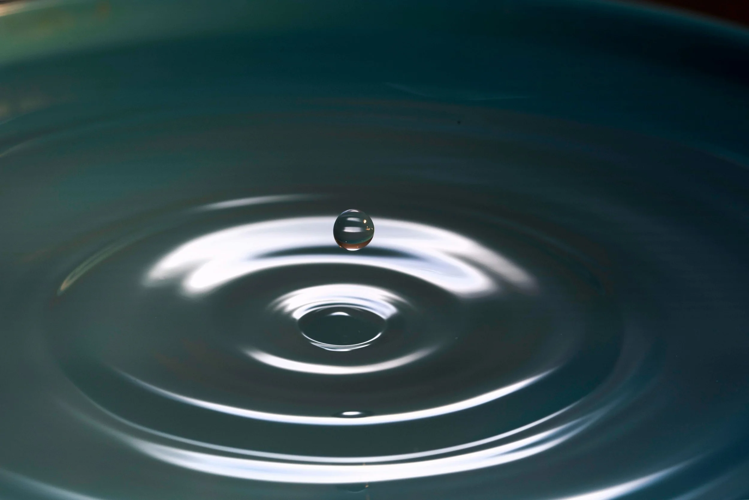 A close-up of a water droplet creating ripples on the surface of a water body, with a dark background.
