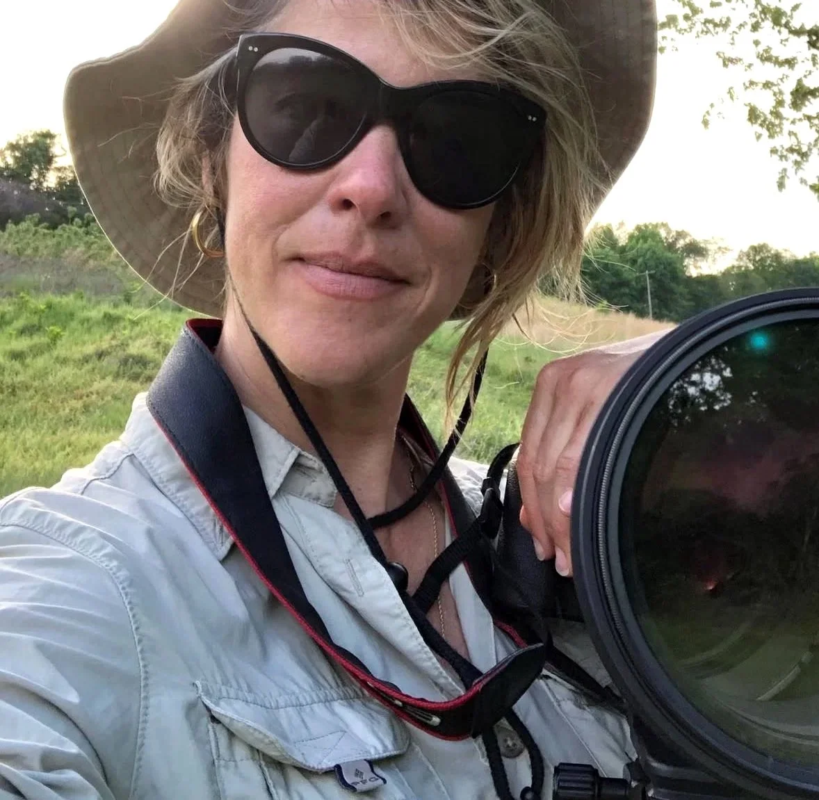 Woman wearing sunglasses and a hat, holding a large camera lens outdoors.