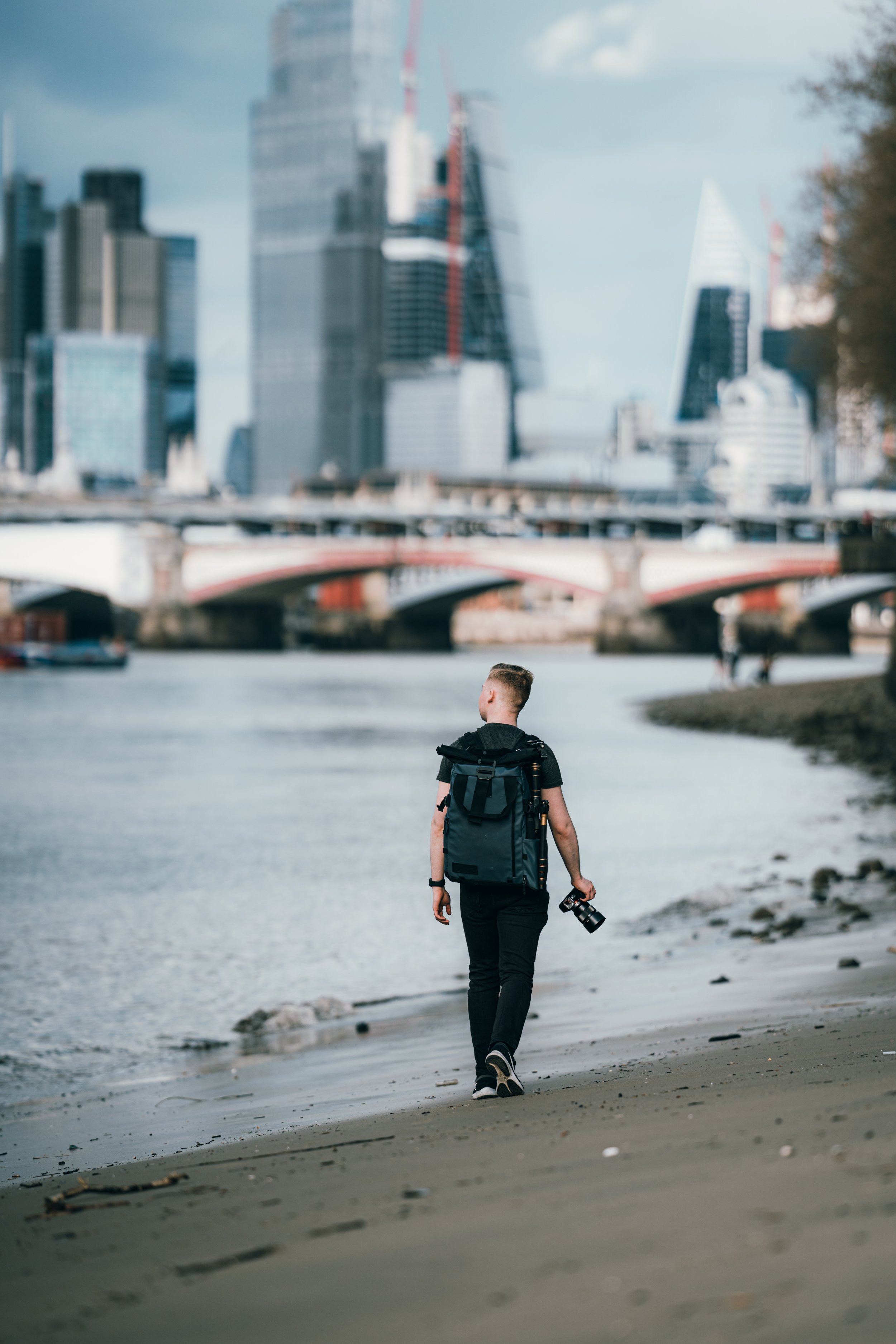 Young person walking along the riverbank with a camera in hand, carrying a backpack, with a city skyline and bridge in the background.