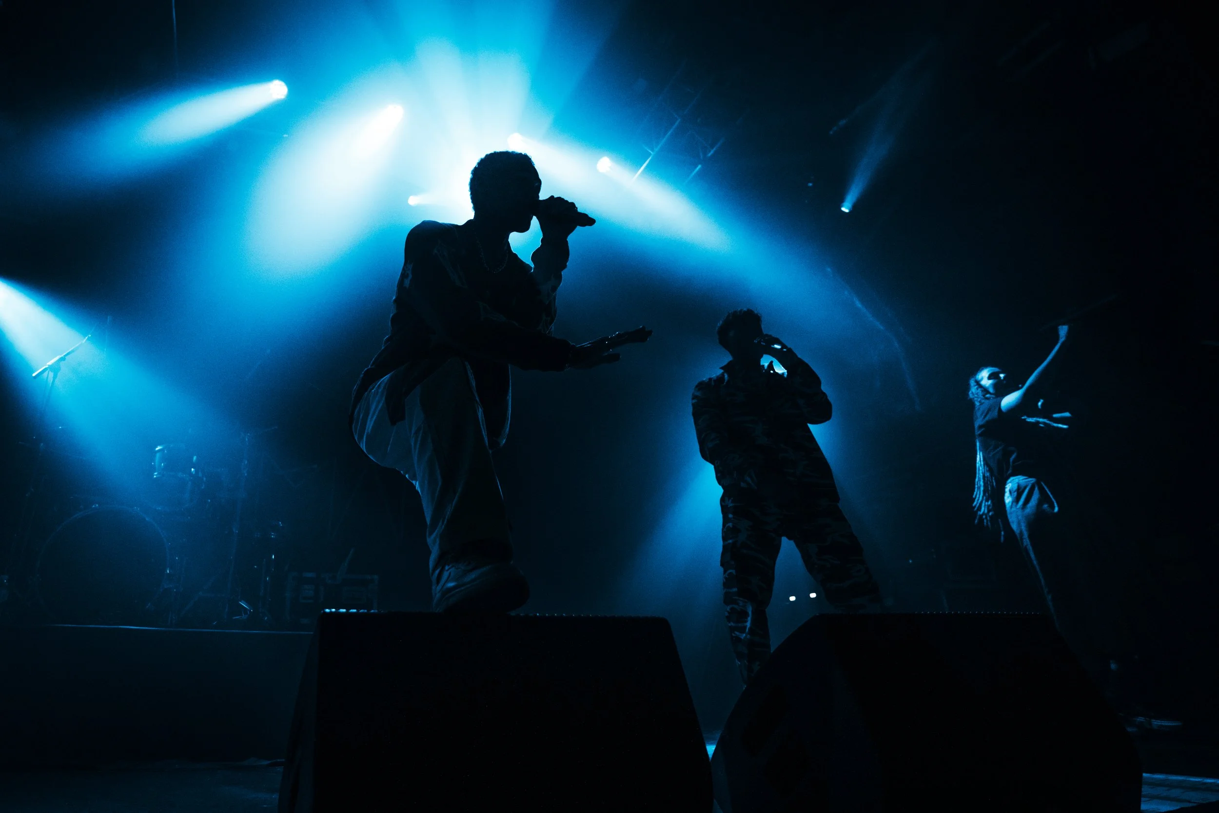 Silhouettes of three performers on stage under blue lighting, holding microphones, with musical equipment in the background.