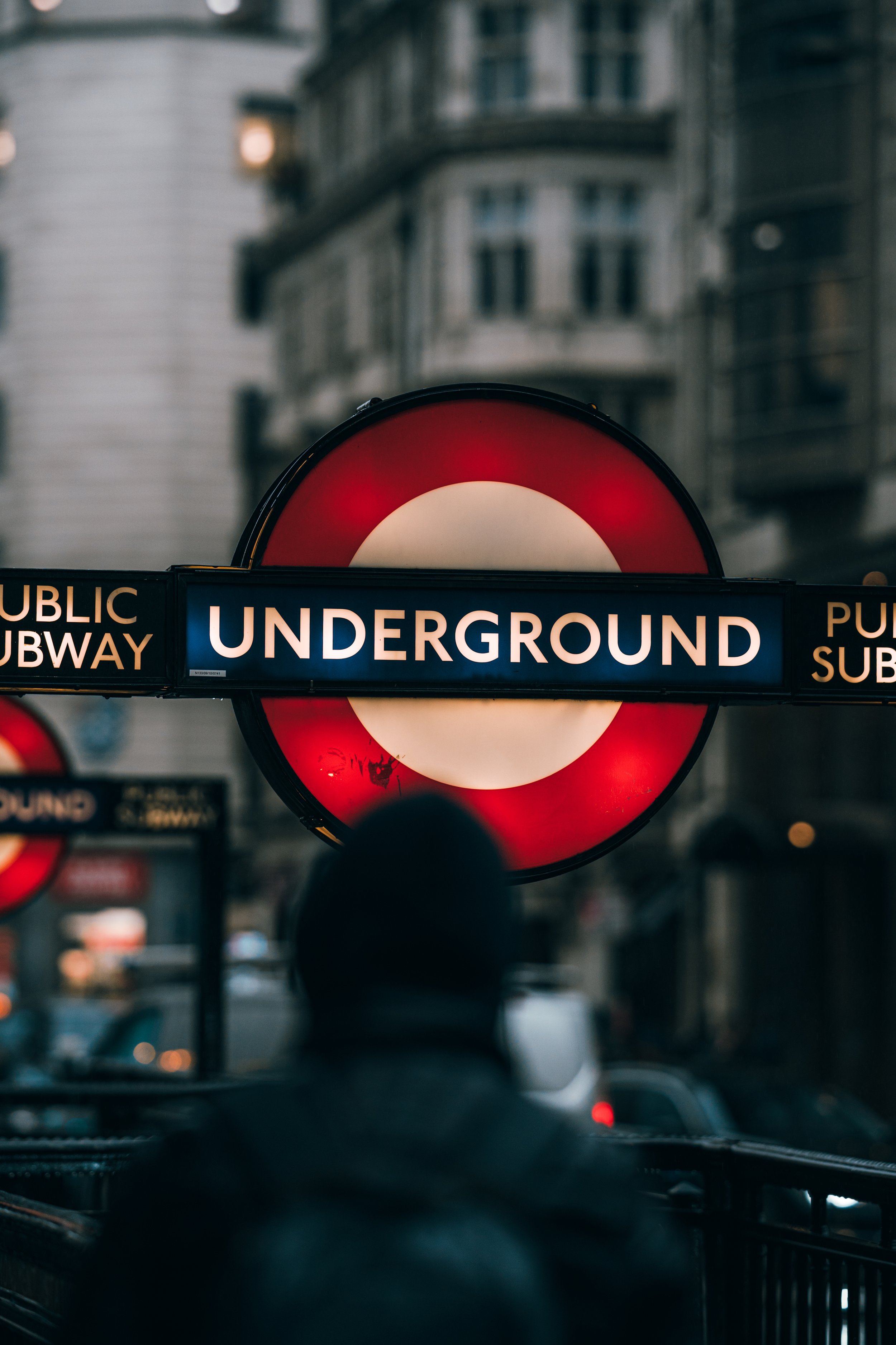 London Underground sign with a person in a dark coat in the foreground, buildings in the background.
