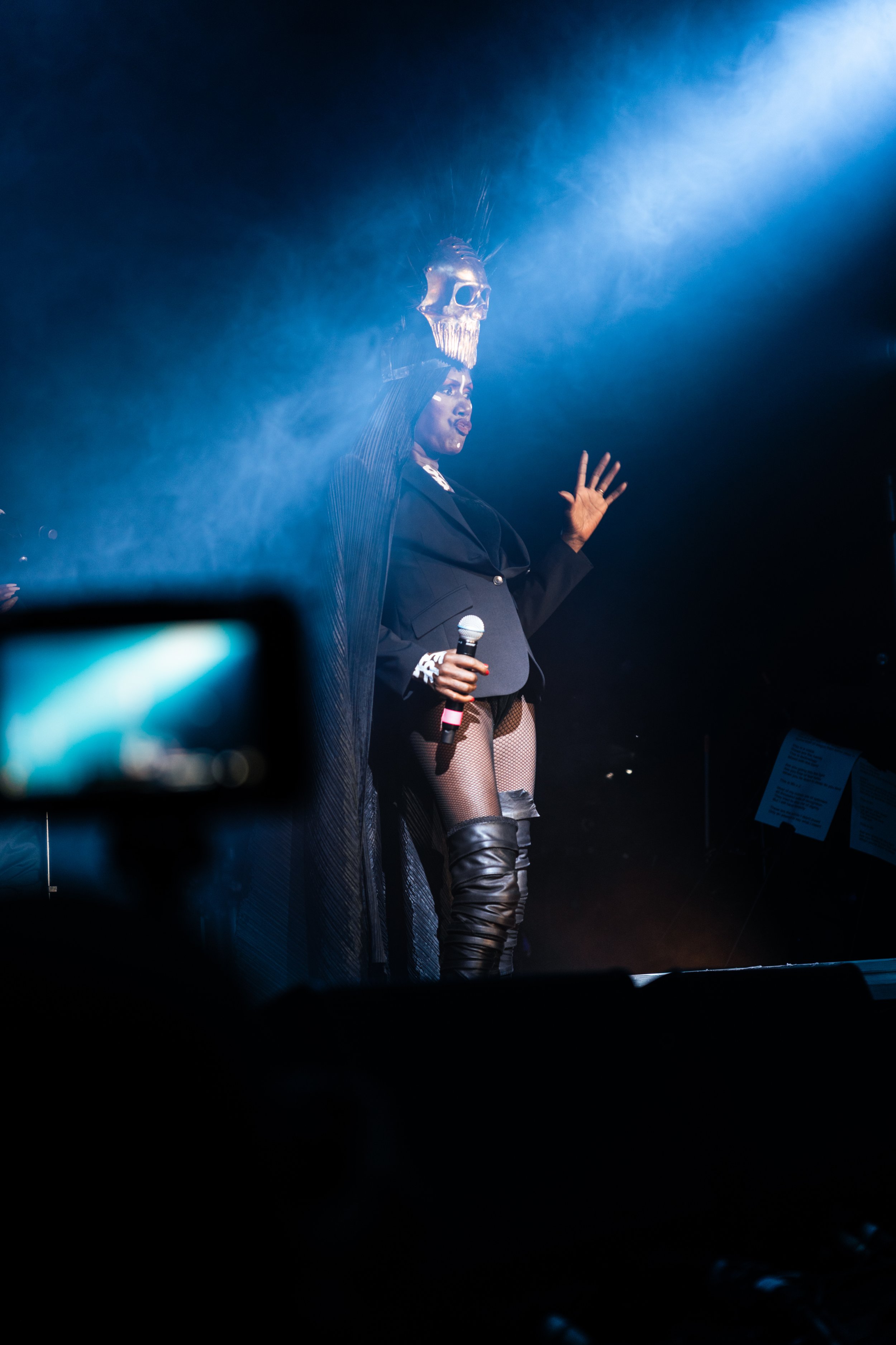 Performer on stage wearing a black blazer, fishnet stockings, and thigh-high leather boots, holding a microphone, with a skull headpiece above her head and dramatic stage lighting.