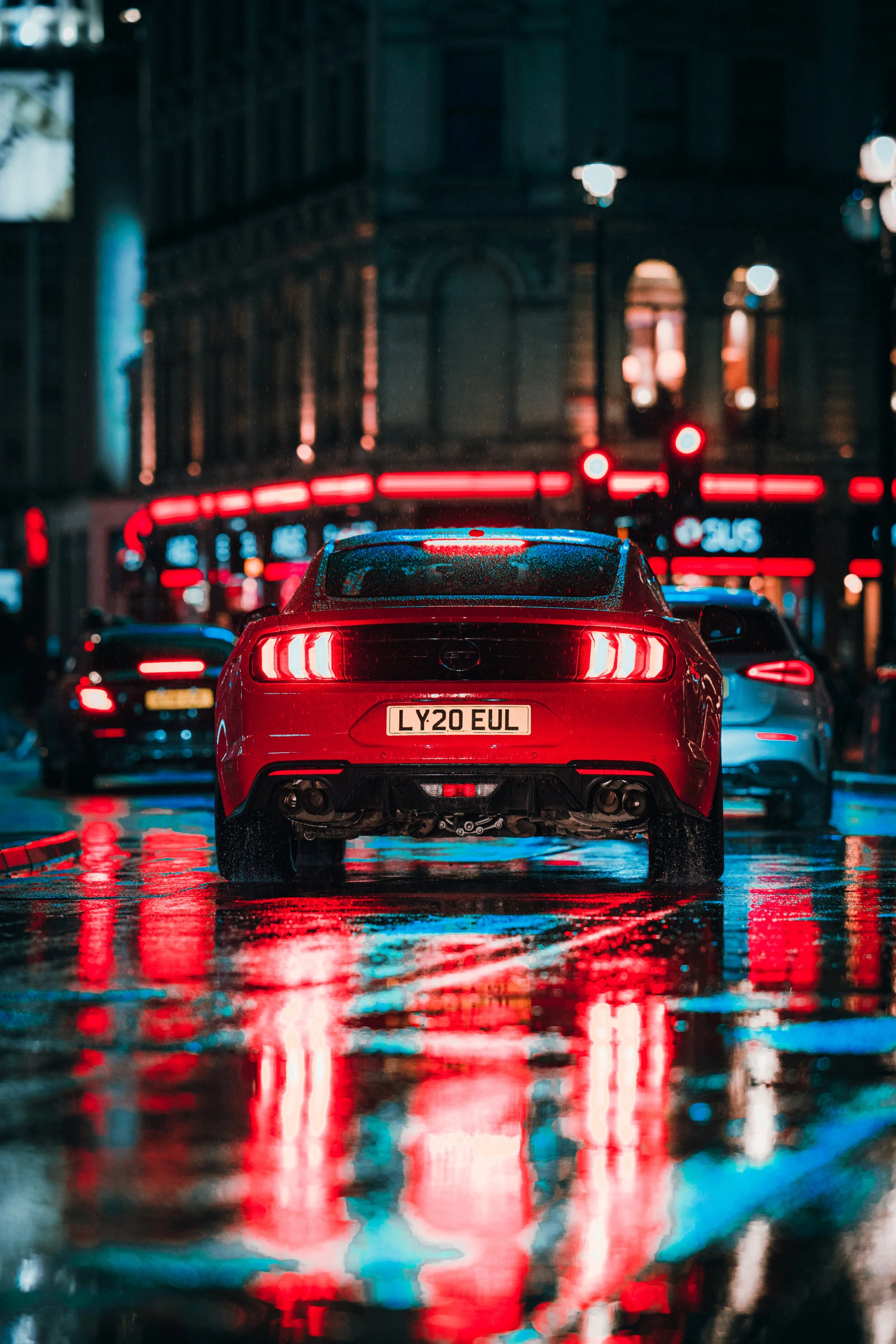 A red sports car with illuminated tail lights is parked on a wet city street at night, reflecting neon lights from surrounding buildings.