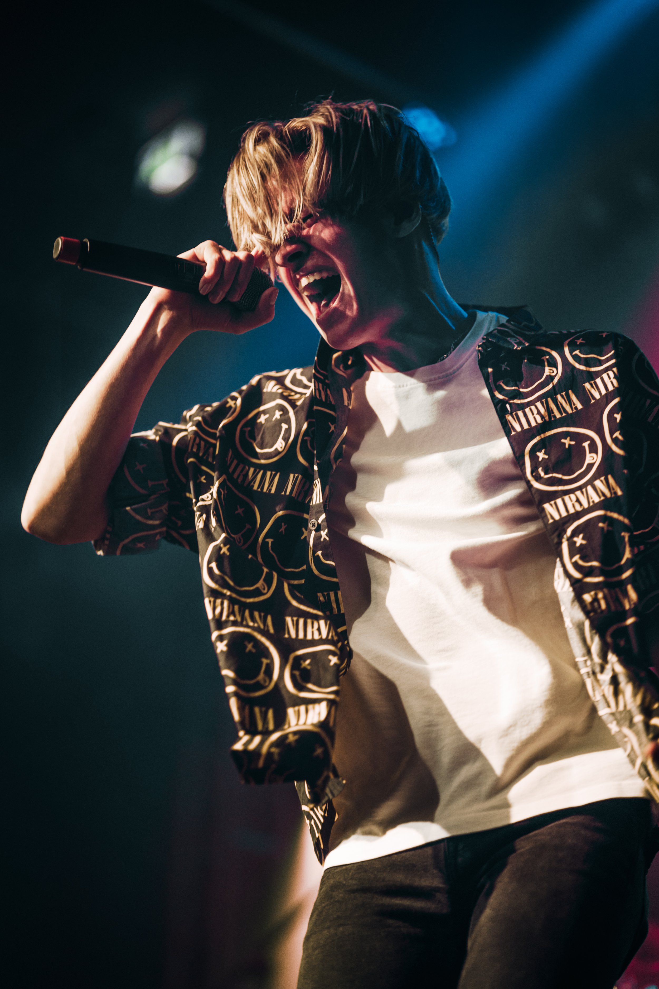 A young person passionately singing or shouting into a microphone on stage, wearing a Nirvana-themed shirt.