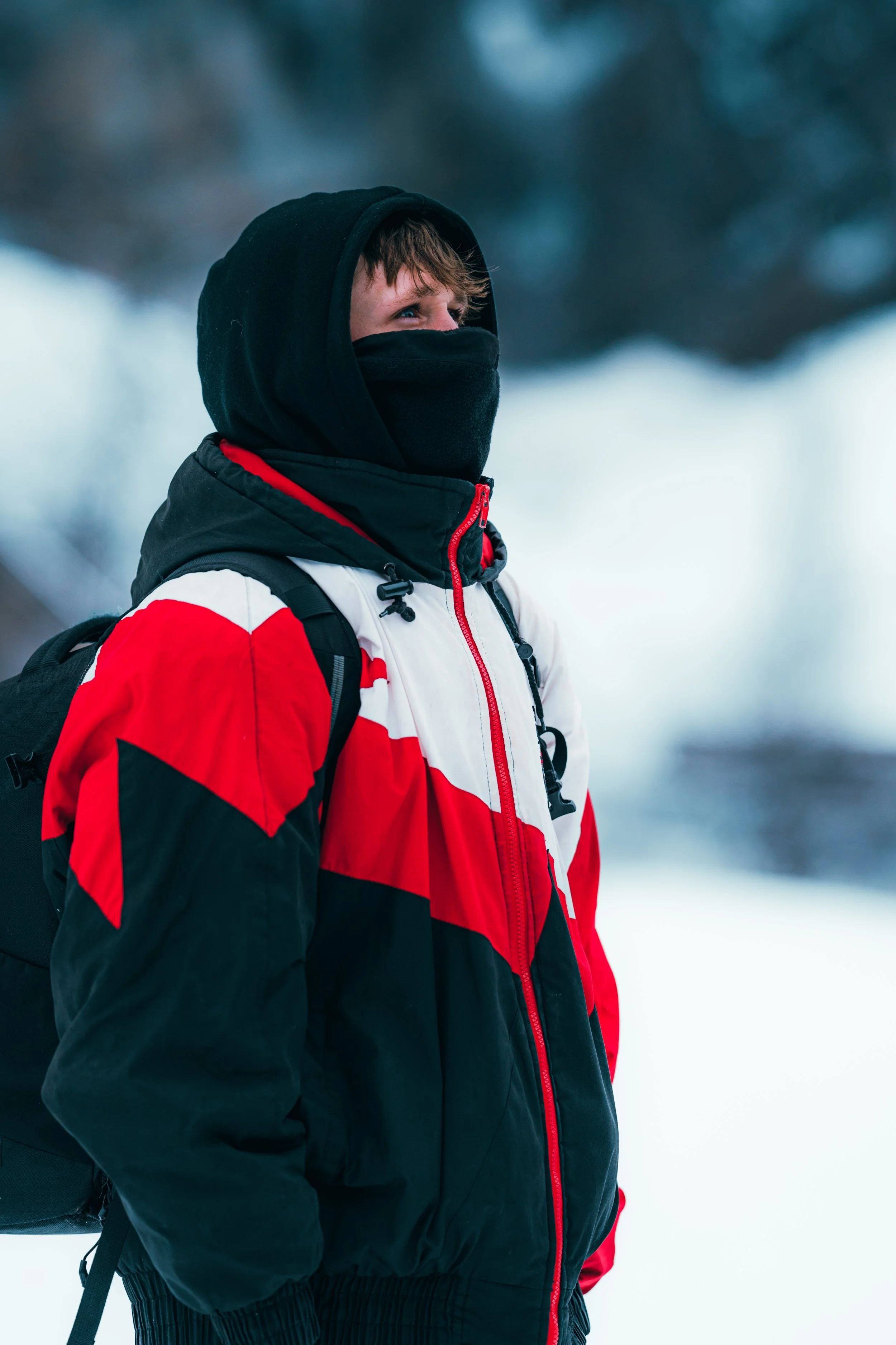 Person dressed in winter gear with a red, white, and black jacket, black face covering, and backpack standing outdoors in a snowy landscape.
