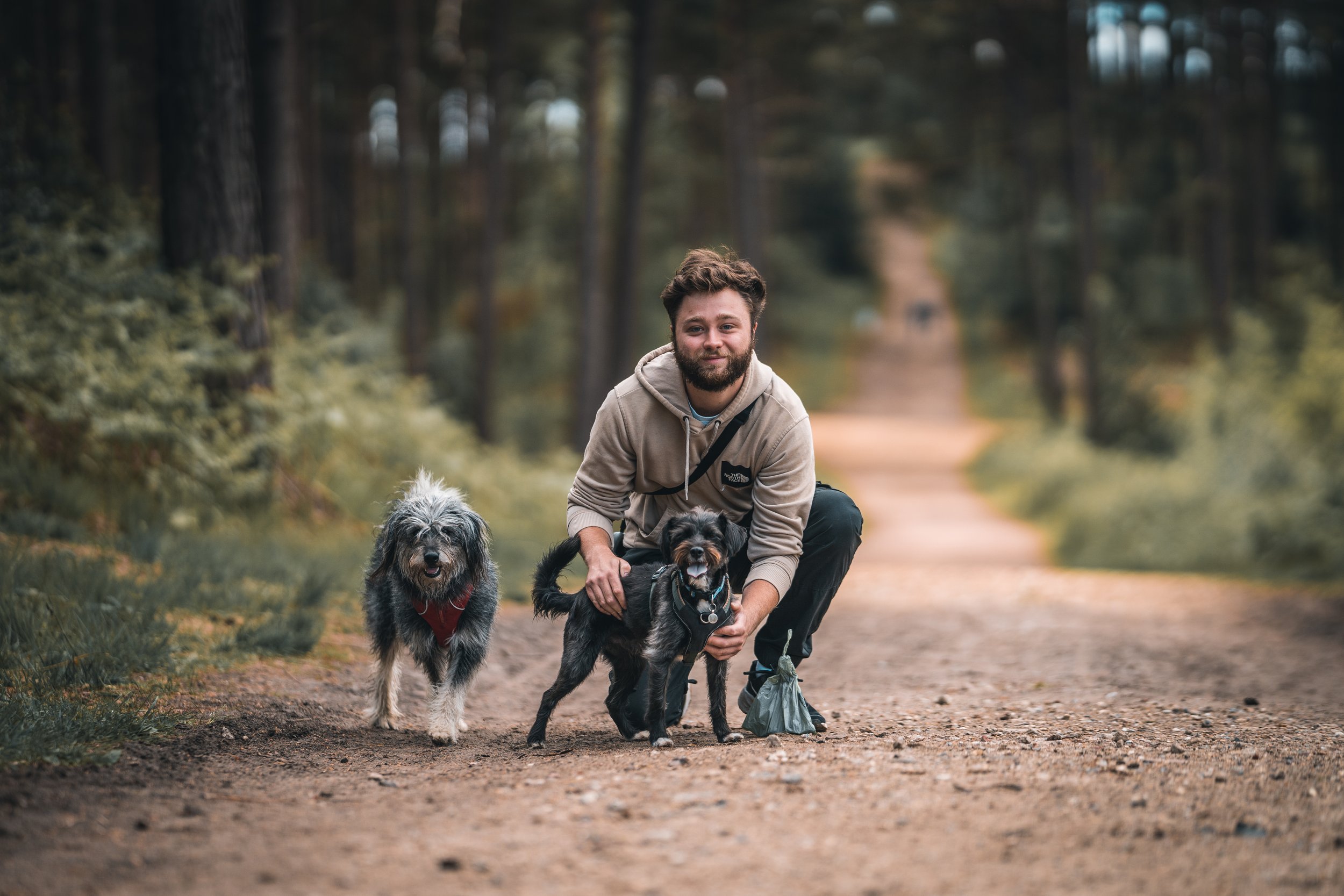 A man with a beard crouching on a dirt trail in a forest, holding two dogs on leashes, with trees lining the trail in the background.