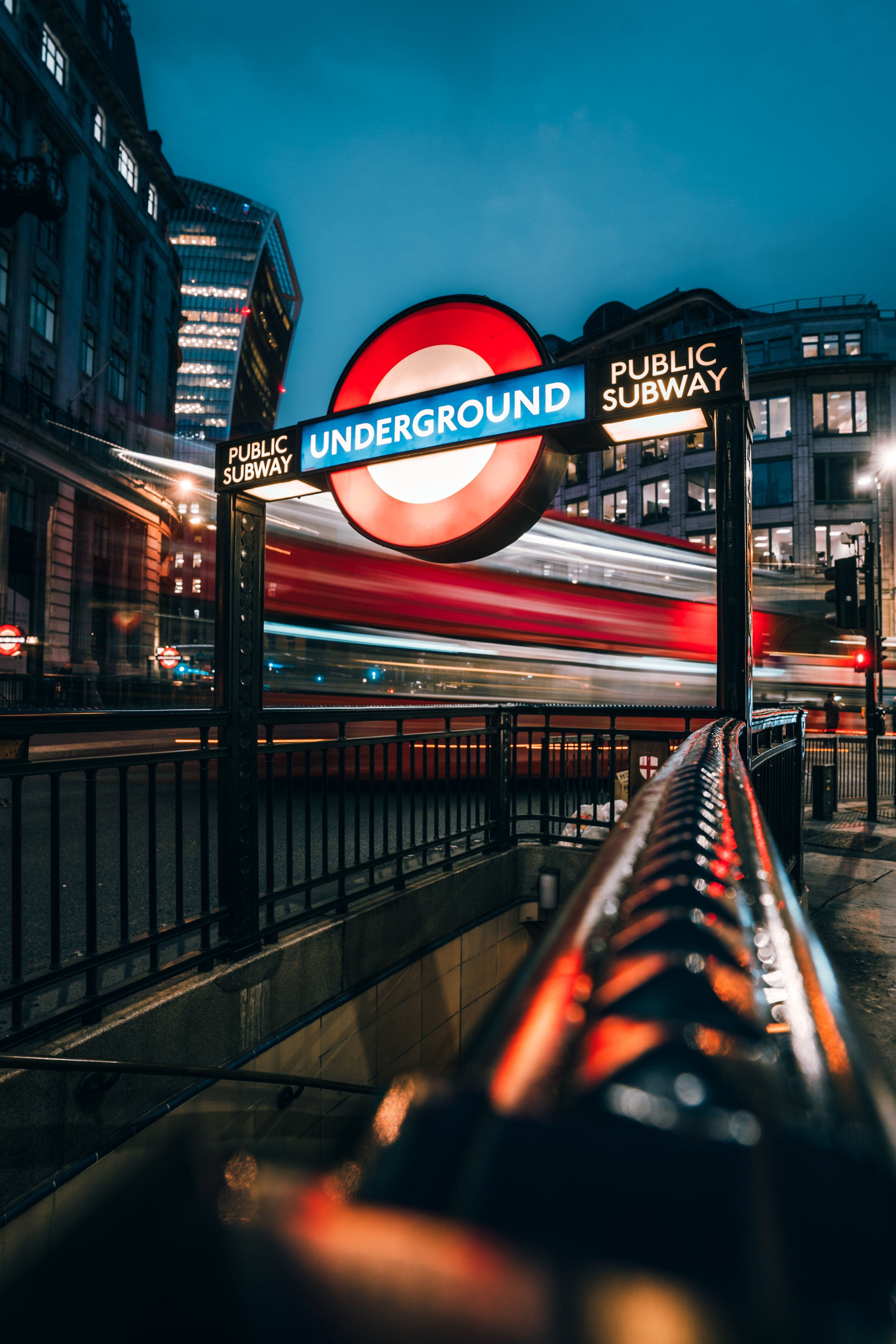 London Underground sign at night with a blurred red double-decker bus passing in the background.