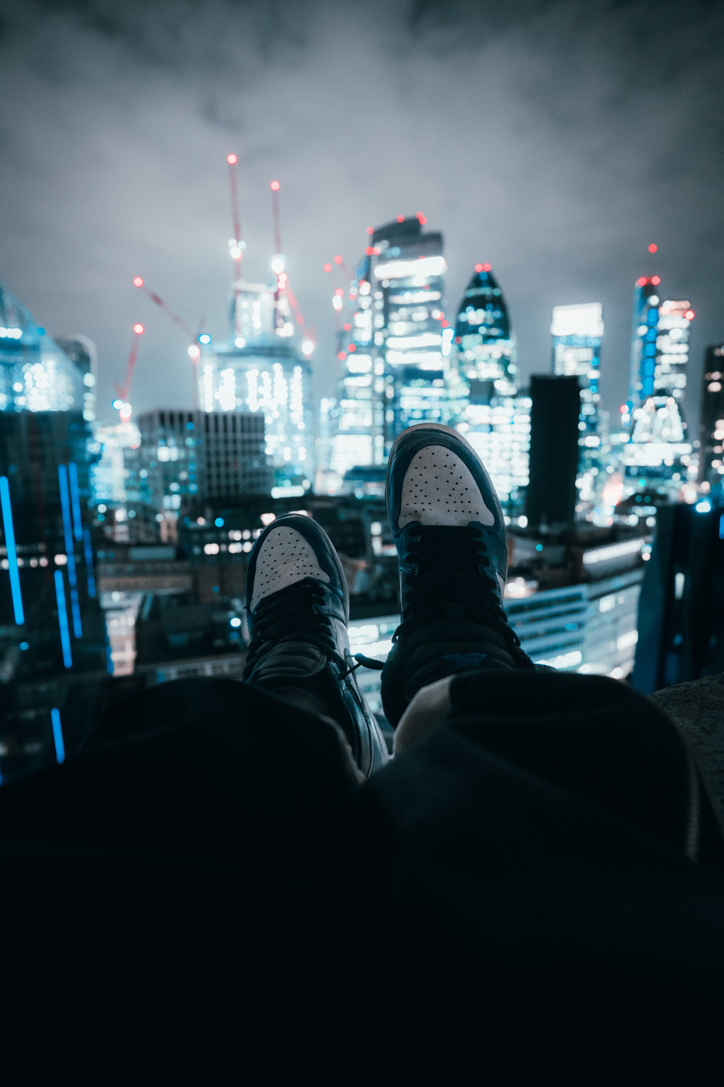A person wearing sneakers sitting on a ledge overlooking a city skyline at night with illuminated skyscrapers and construction cranes.