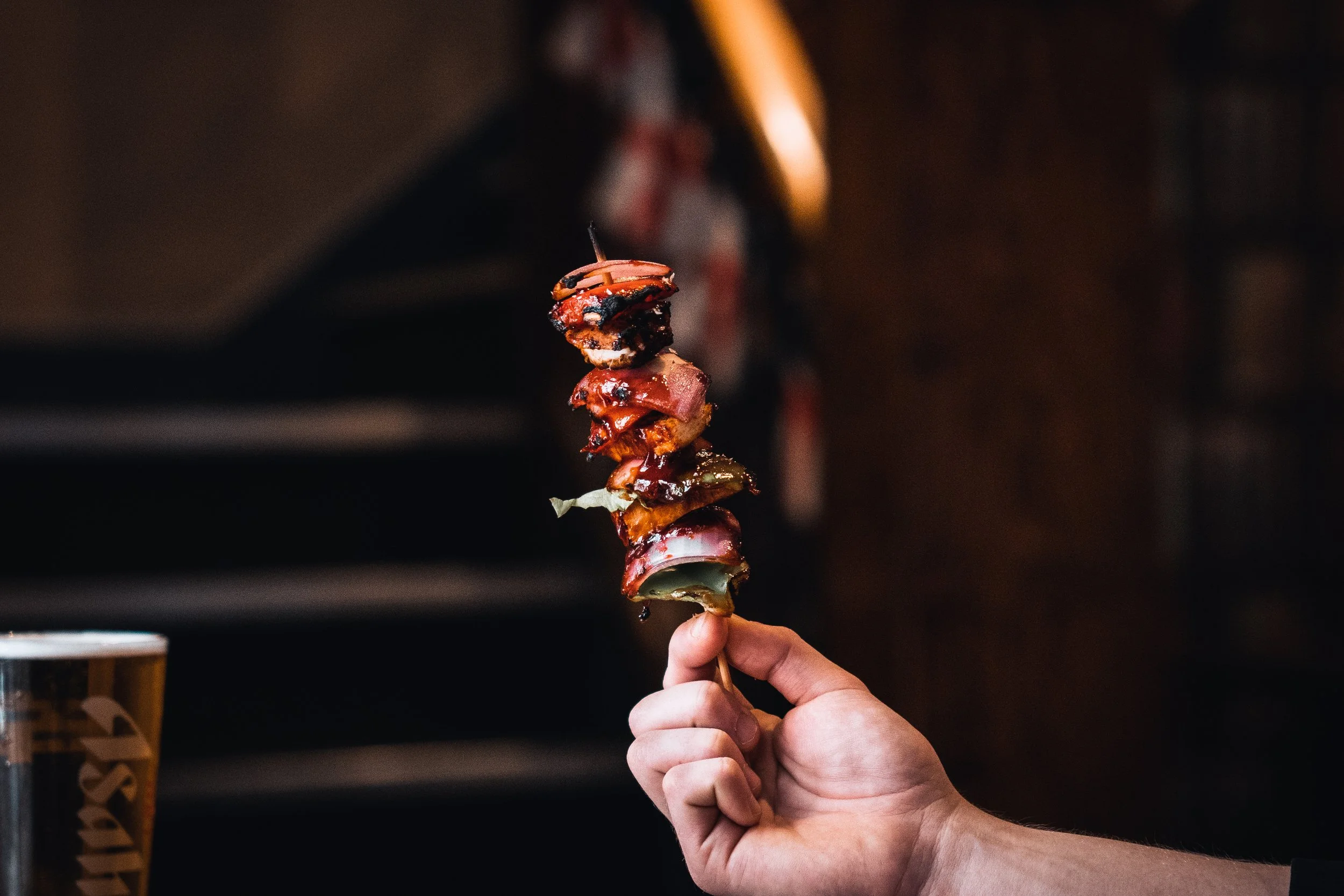 A hand holding a skewer with grilled meat and vegetables, with a dark, blurred background and a glass of beer to the left.