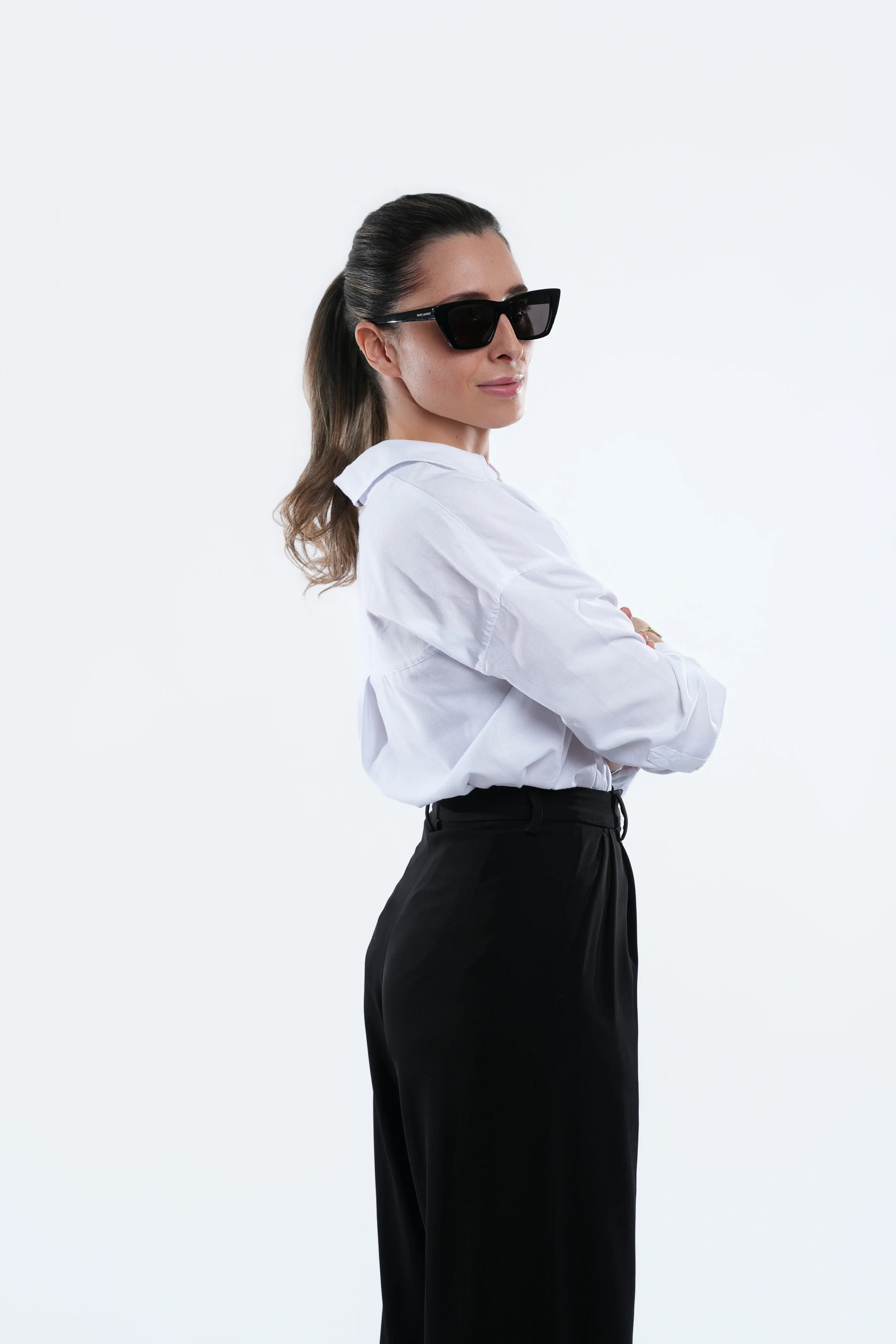 Woman in white shirt and black trousers standing sideways with arms crossed and sunglasses, fashion portrait New Moon Agency Klagenfurt
