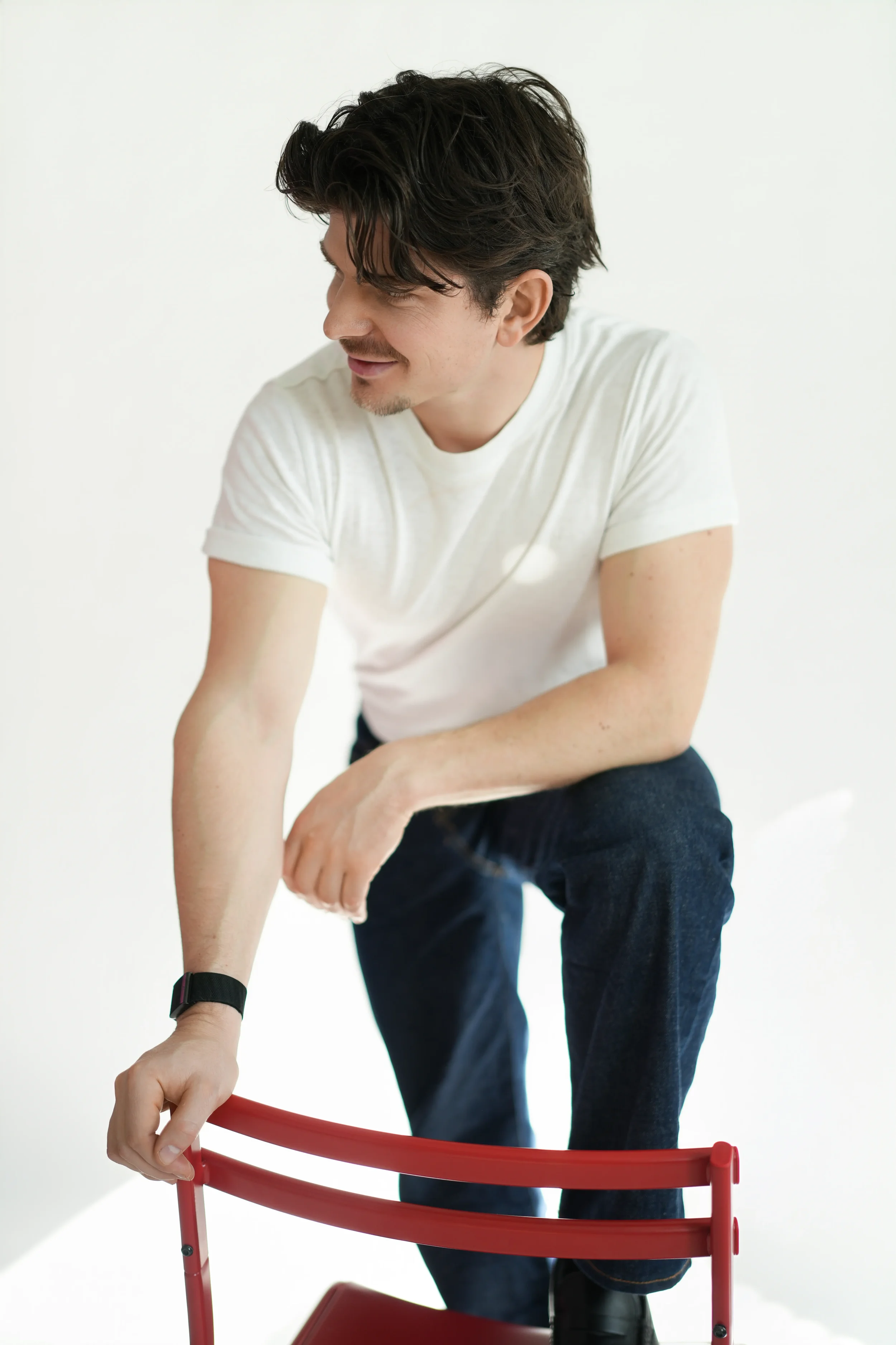 Man in white t-shirt sitting on red chair looking away with a smile, relaxed studio portrait New Moon Agency Klagenfurt