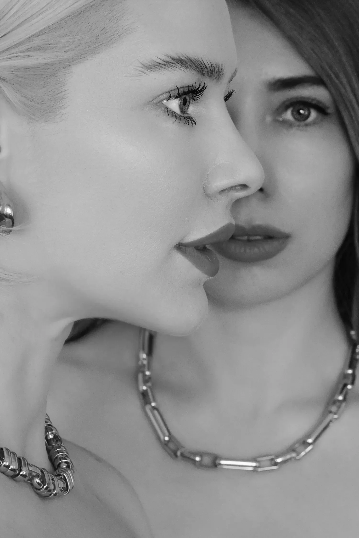 Nahaufnahme eines Schwarz-Weiß-Porträts von zwei Frauen: Die eine hat helle Haut und blonde Haare, die andere dunklere Haare; beide tragen metallischen Schmuck und blicken sich mit ernsten Mienen an.