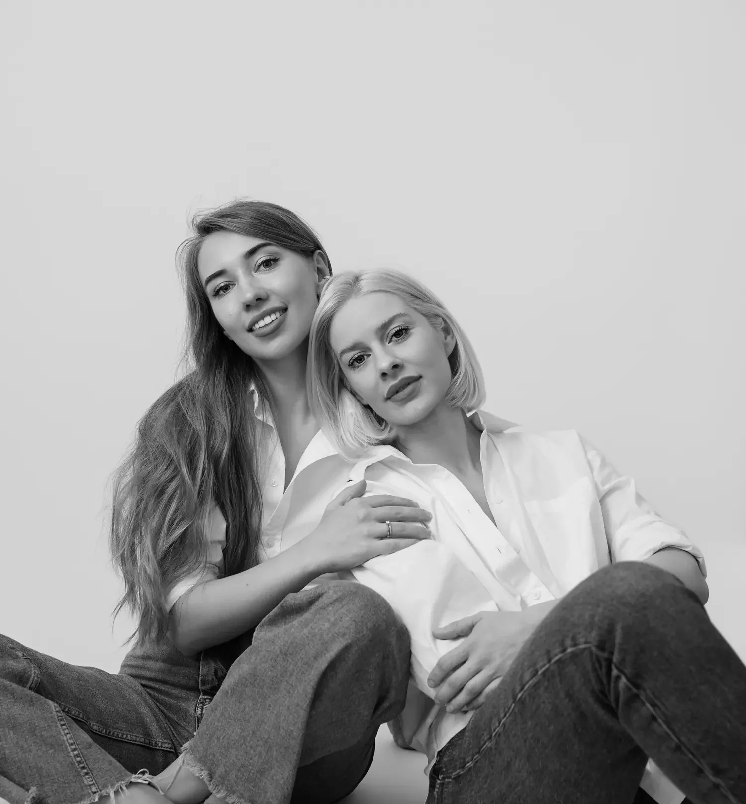 Zwei Frauen, die dicht beieinander sitzen, die eine mit langen, welligen Haaren, die andere mit kurzen, glatten Haaren; beide tragen weiße Hemden und Jeans und lächeln auf einem Schwarz-Weiß-Foto in die Kamera.