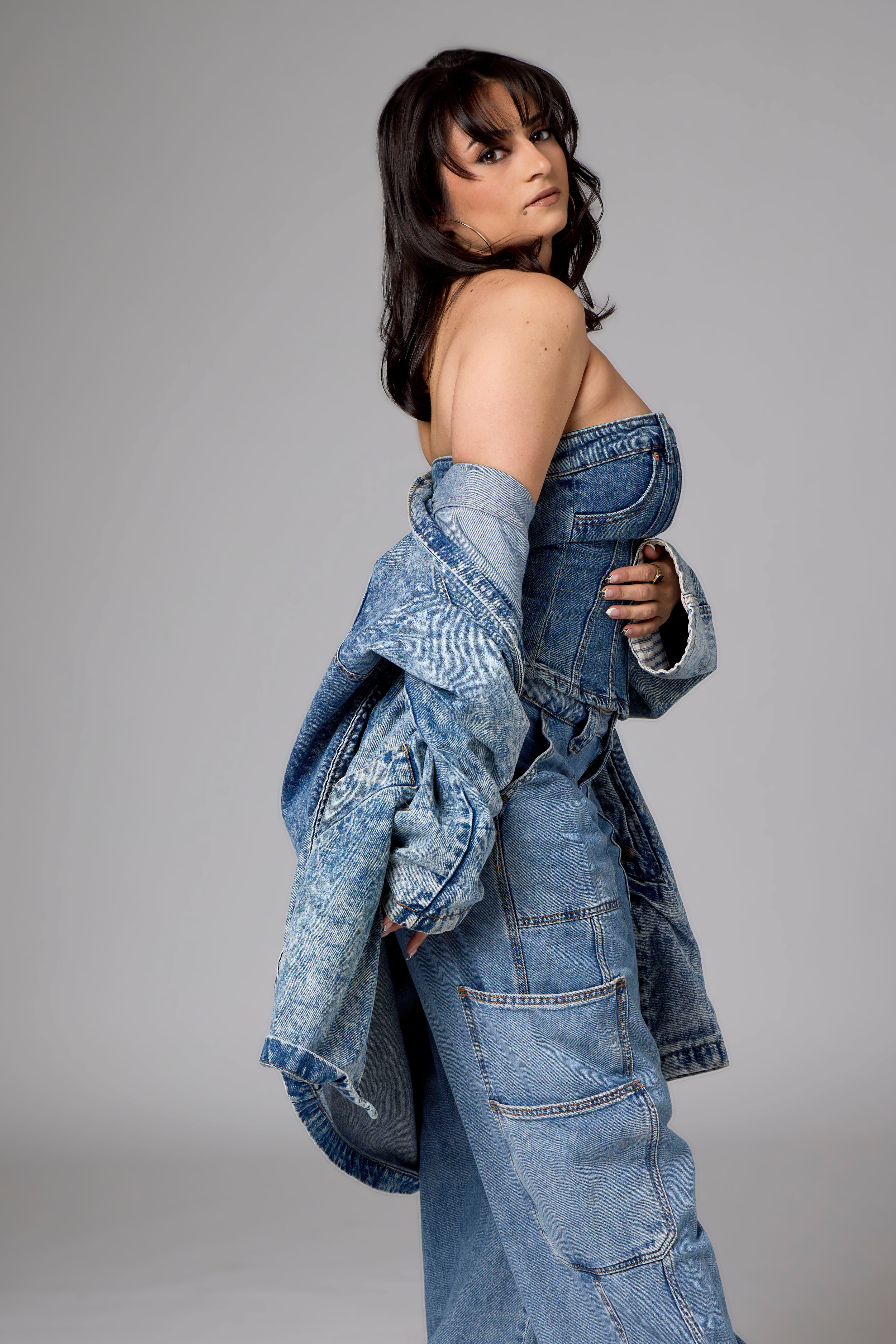 A young woman with dark brown hair styled in waves wearing a denim jacket draped over her arms, a denim bustier top, and high-waisted jeans with multiple pockets, standing against a plain gray background.