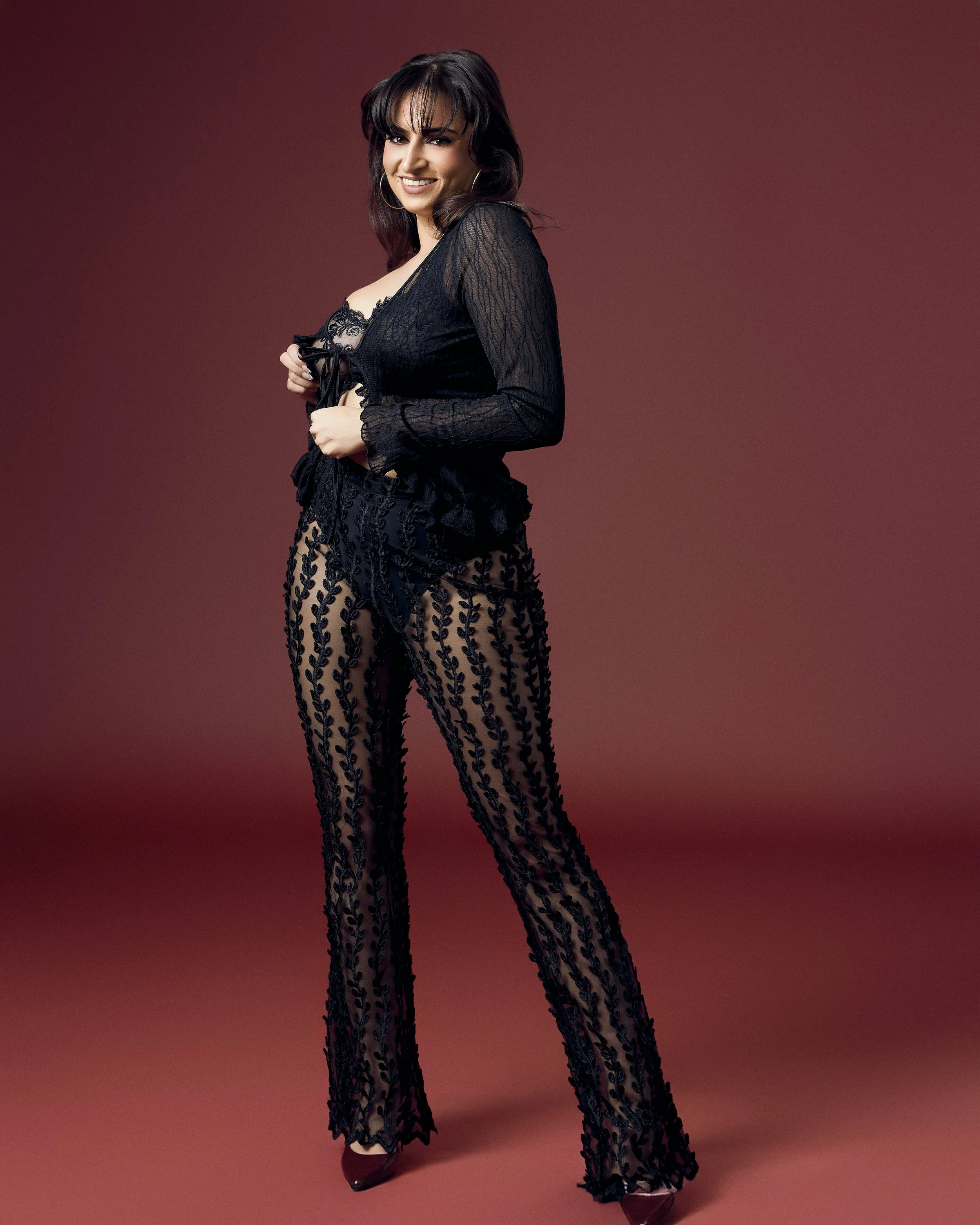 woman in black lace sheer top and pants with embroidered leaf pattern, posing against a red gradient background