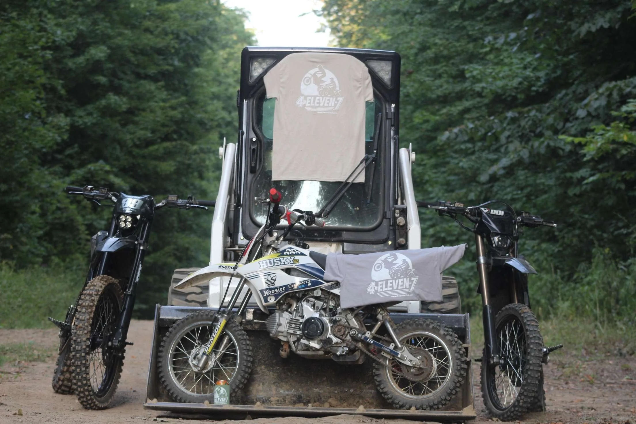 A dirt bike in front of a utility vehicle, with matching T-shirts draped over the bike and vehicle that have a 4-Eleven-7 logo, with green trees in the background.