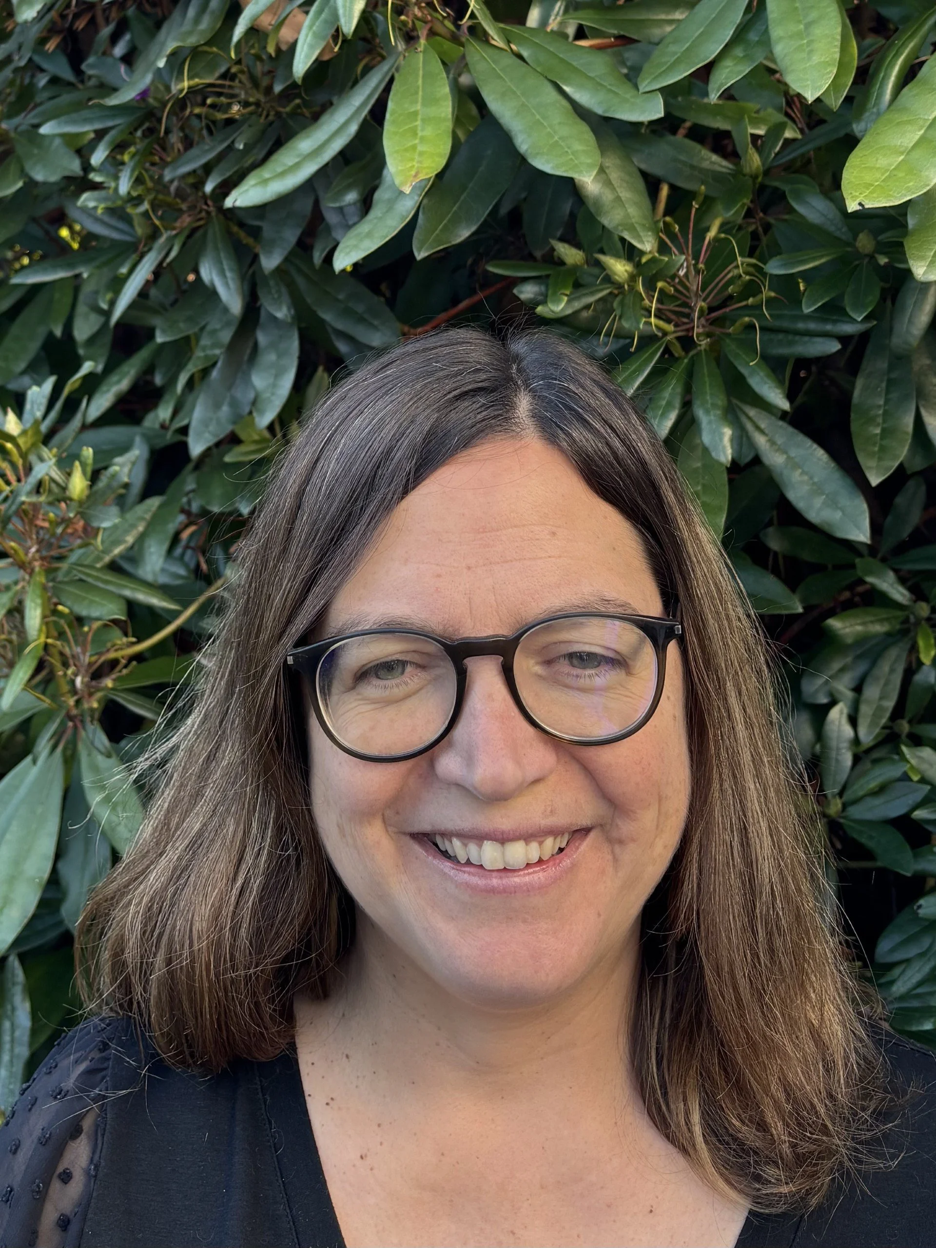 Dr Debbie Cooke, a woman with shoulder-length brown hair, wearing glasses, smiling, standing outdoors in front of green leafy bushes.