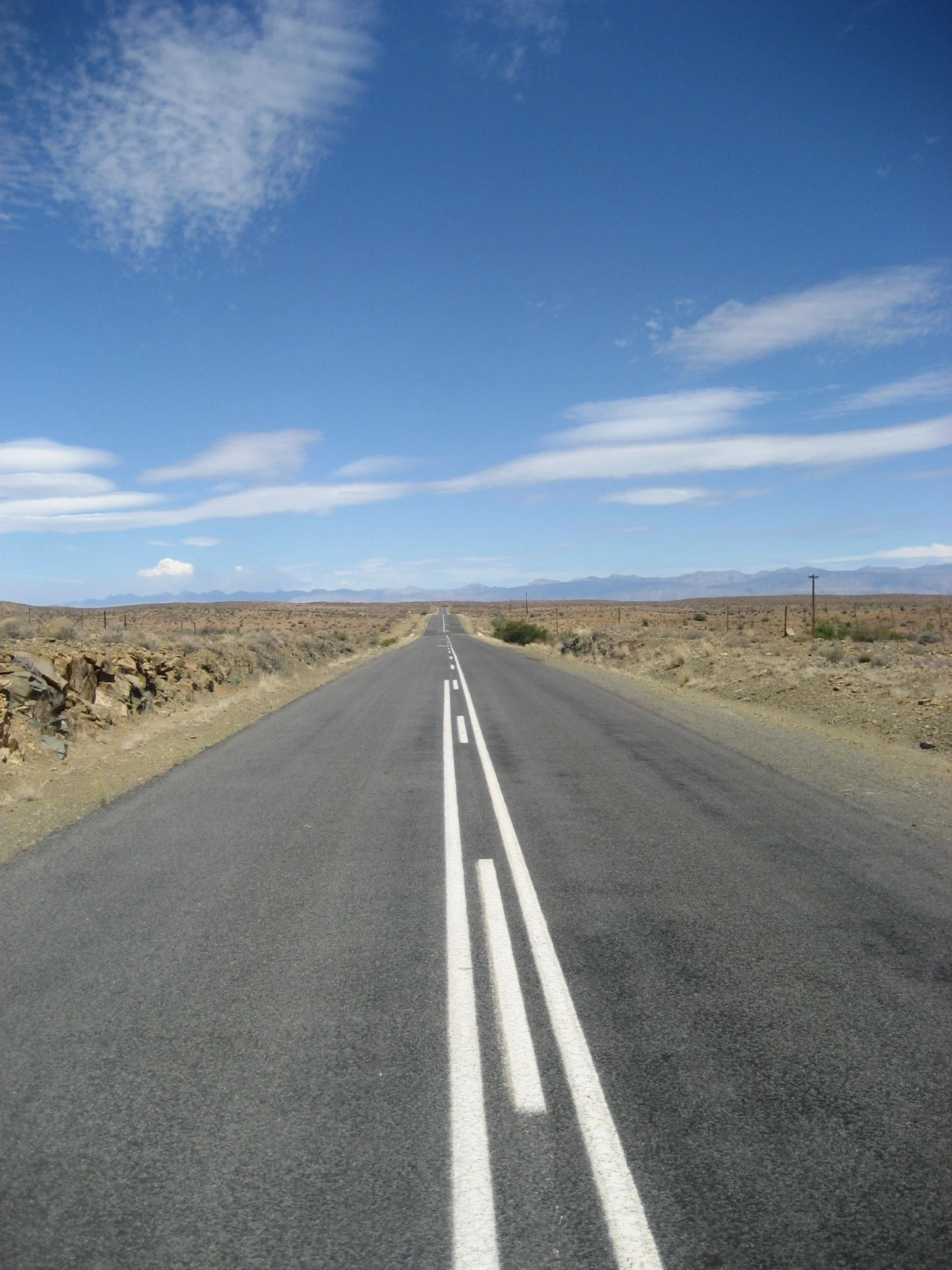 A straight, two-lane road in a desert landscape under a blue sky with clouds, distant mountains on the horizon.