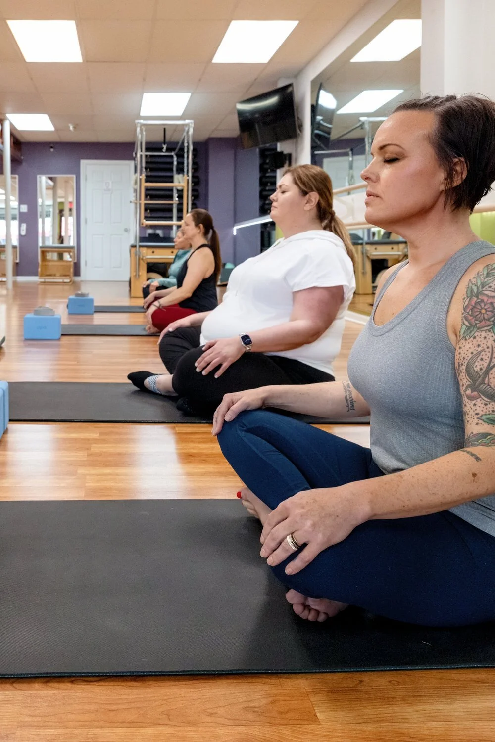 Women practicing yoga or meditation in MBS Pilates fitness studio, sitting cross-legged on yoga mats with focused expressions.