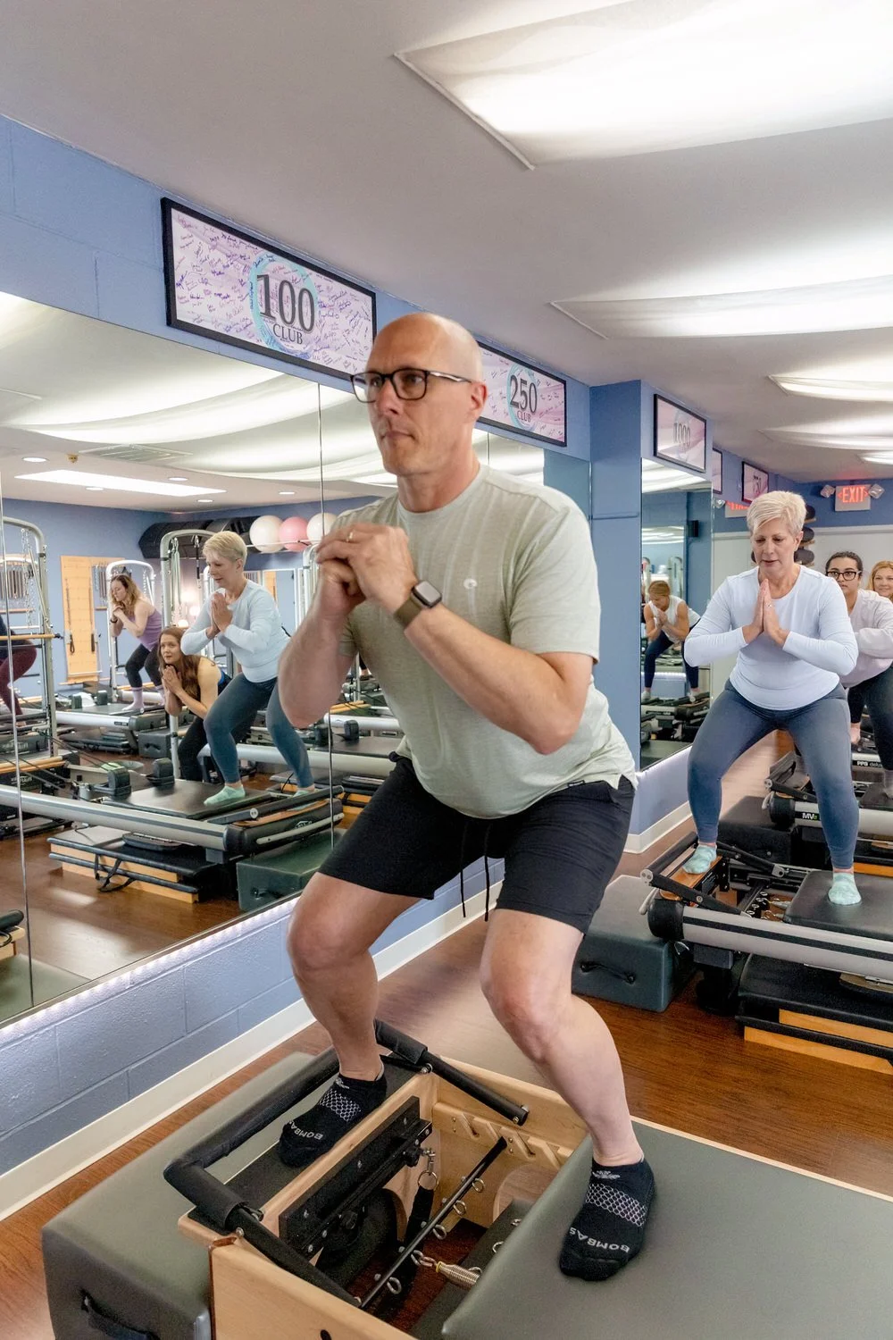 A man on a Pilates reformer machine in a fitness class, surrounded by mirrors and other participants, with a mirror showing signs for different club levels in the background.