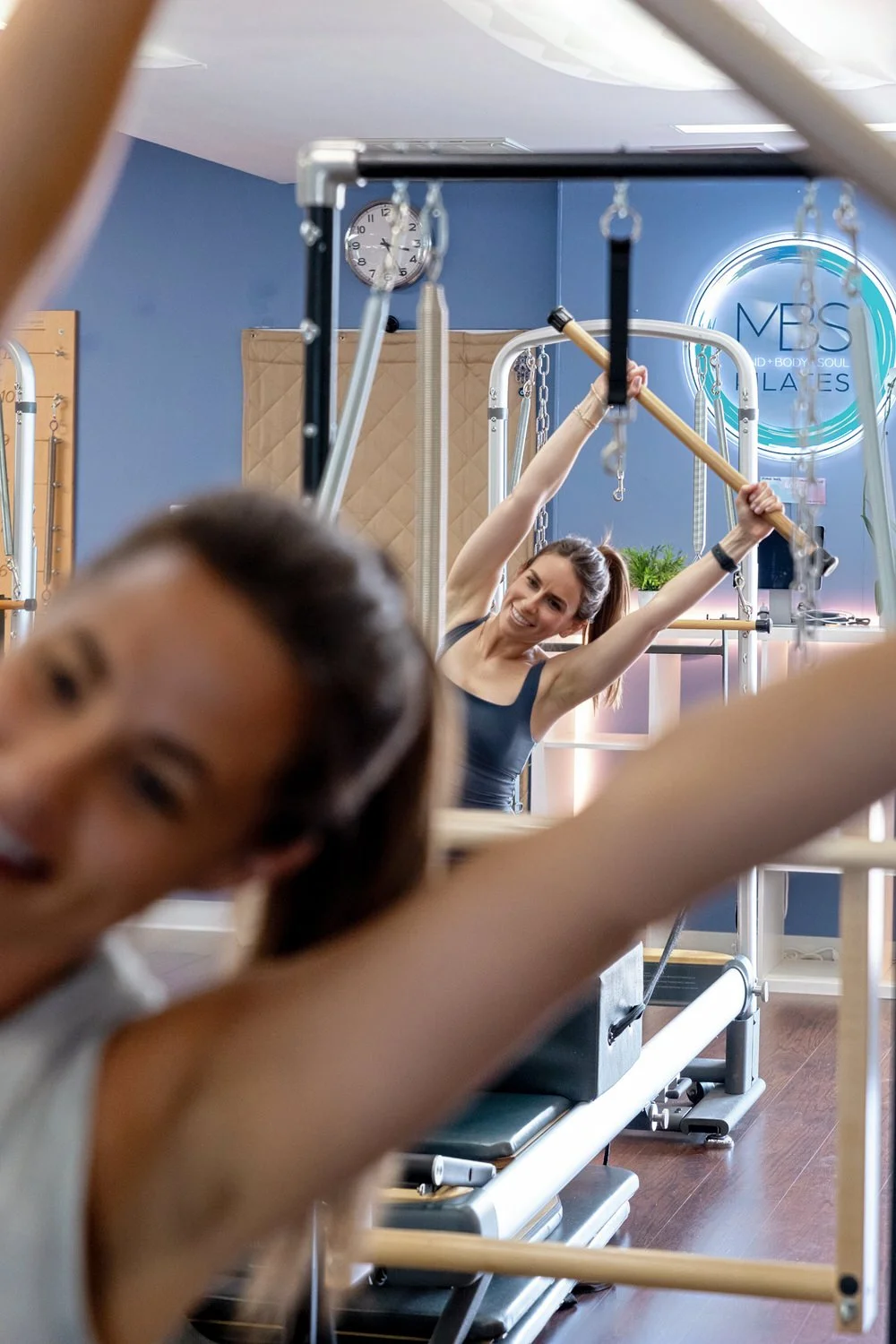 Two women exercising with Pilates equipment at MBS Pilates, smiling and stretching, with a blue wall and fitness branding in the background.