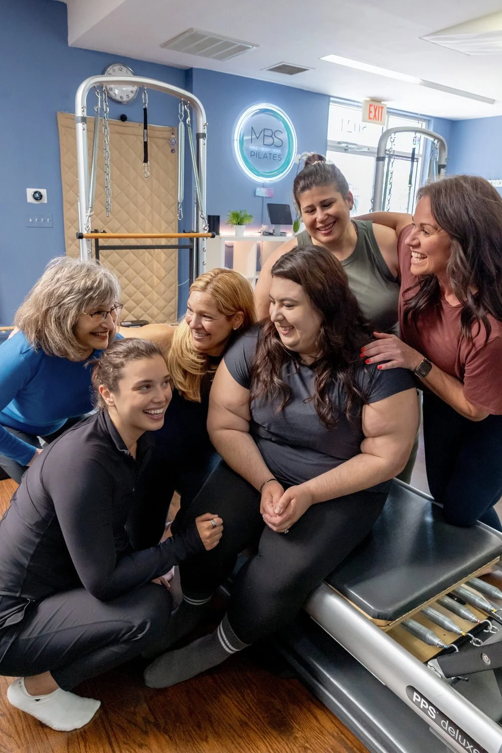 Six women of diverse ages and ethnicities smiling and laughing together in MBS Pilates, with one sitting on a Pilates reformer machine and the others gathered around her.