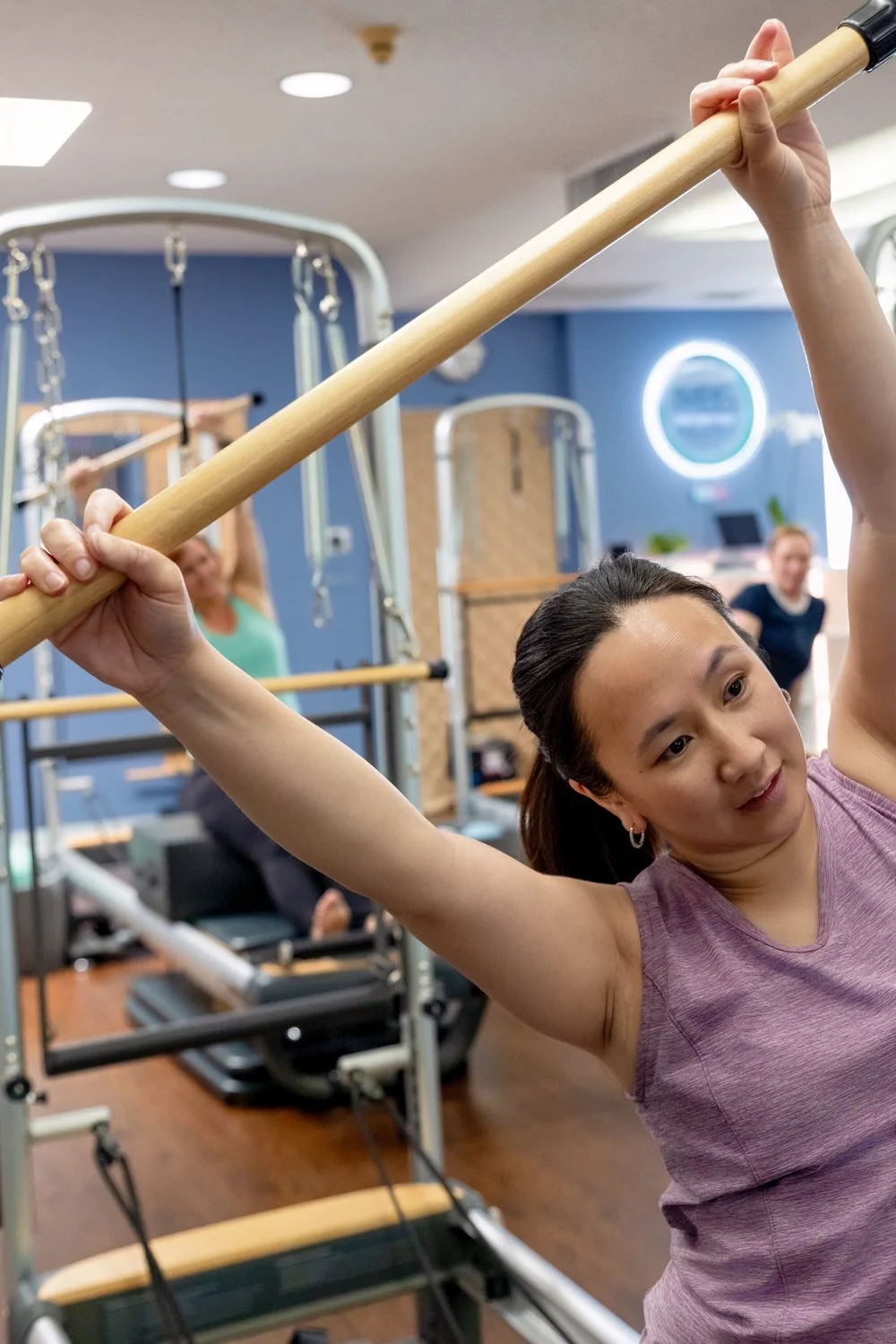 A woman performs a stretching exercise with a wooden pole at MBS Pilates, with other people exercising in the background.