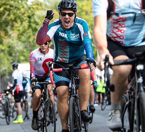 Cyclists participating in a race, with one rider in the foreground celebrating victory, wearing a blue and red jersey, helmet, and sunglasses, during daytime.