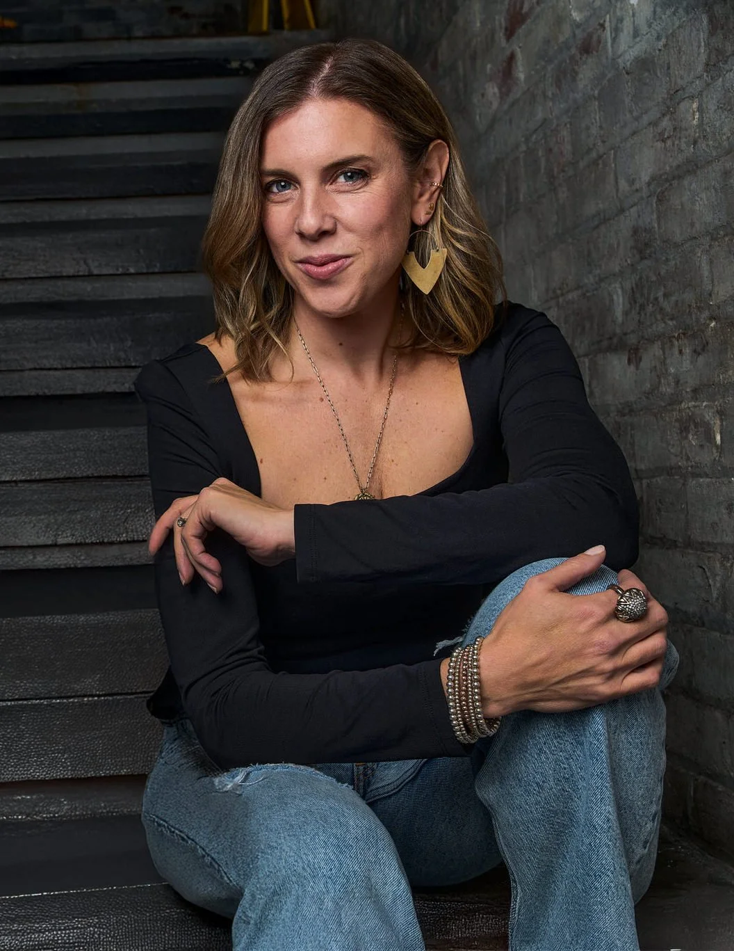 A woman with shoulder-length brown hair wearing a black long-sleeve top, ripped blue jeans, and jewelry including large earrings, rings, bracelets, and a necklace. She is sitting on stairs with a brick wall in the background, smiling and looking at the camera.